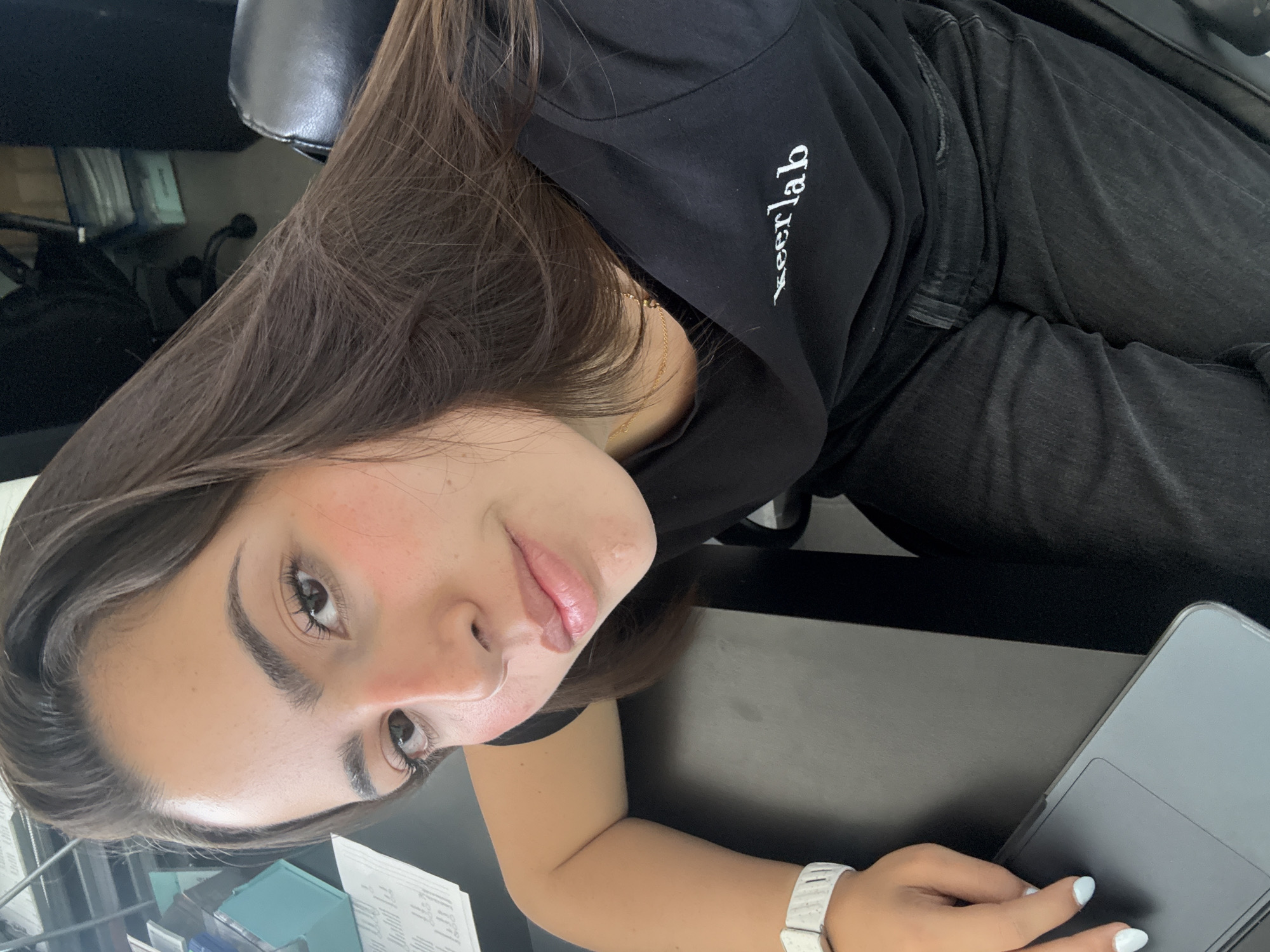 A young woman is captured in an angled, close-up indoor shot, likely in San Bernardino Tlaxcalancingo, Mexico. She has long, dark brown hair that cascades over her shoulder, fair skin, and a soft expression with a faint smile. Her eyes are light brown, and she wears subtle makeup with noticeable blush on her cheeks and light pink lipstick. She is dressed in a black hoodie that features the white, stylized text "Keerlab" on the front, paired with dark-colored jeans. A delicate gold chain is visible around her neck. Her left arm rests on a dark-colored laptop or large tablet, revealing a white textured wristband or watch and light blue nail polish on her fingertips. The setting appears to be a workspace, with a black desk and part of a black office chair visible. To her left on the desk, there are some papers and small, clear containers. The lighting suggests an indoor environment.