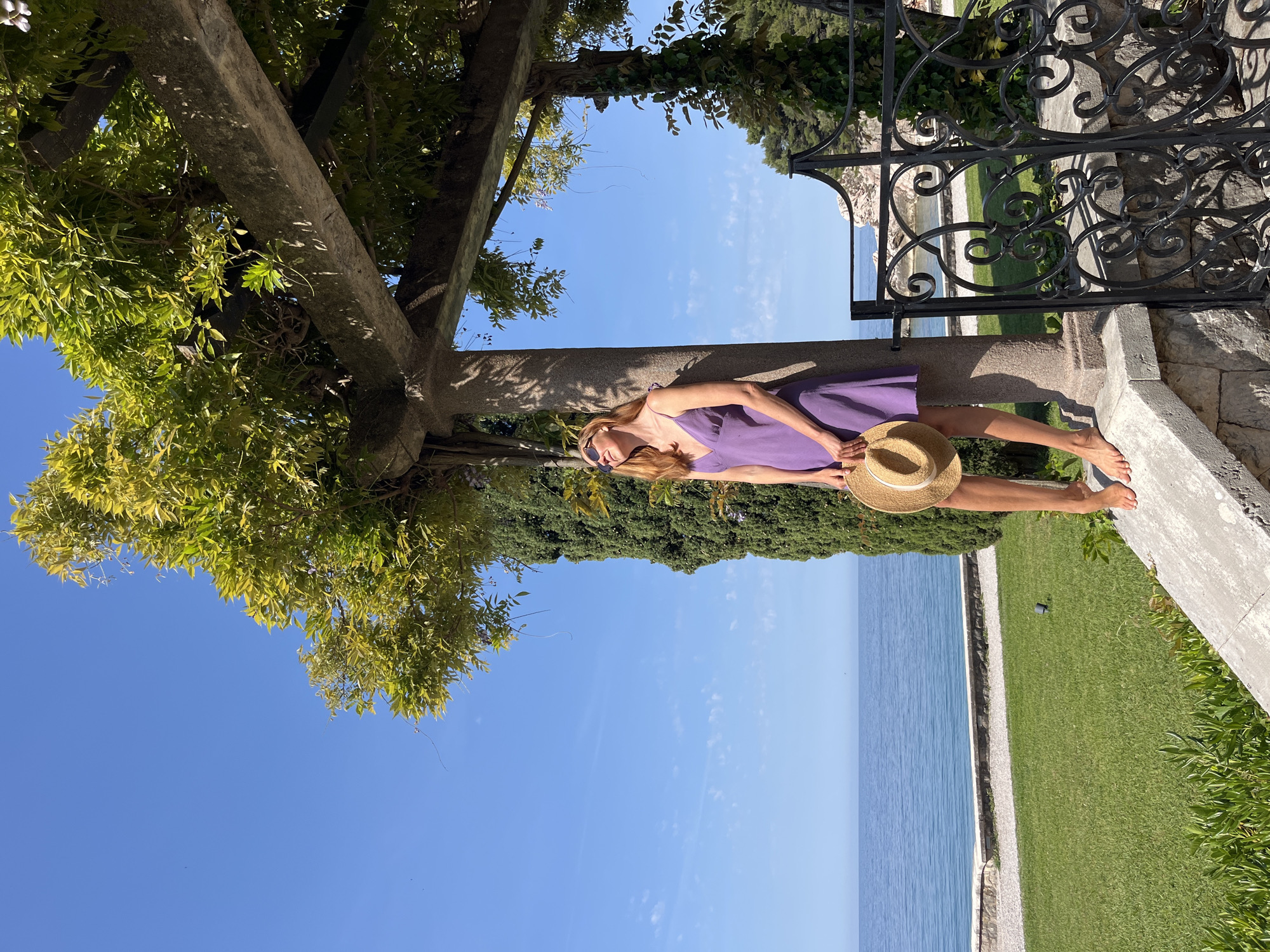 A woman with long, reddish-blonde hair, wearing a purple sundress and sunglasses, is captured relaxing outdoors in a beautiful coastal setting in Sveti Stefan, Montenegro. She is reclining on a weathered stone ledge or beam, which is part of a sturdy pergola structure covered in lush green vines and foliage, casting dappled shadows. She holds a straw hat in her lap and is barefoot, revealing painted toenails.
The background offers a picturesque panoramic view. To her left, vibrant green leaves and branches of trees or vines frame the scene from above. To her right, an ornate black wrought iron fence or gate with intricate scrollwork stands as a decorative boundary. Beyond the fence, a manicured green lawn slopes gently towards the calm, deep blue sea. The clear horizon line separates the tranquil water from a bright, expansive blue sky dotted with a few faint, wispy clouds.
The image conveys a sense of peaceful leisure on a bright, sunny day, typical of a Mediterranean climate. The strong sunlight is evident from the clear definition of shadows and the vivid colors of the natural surroundings. No visible text is present in the image.