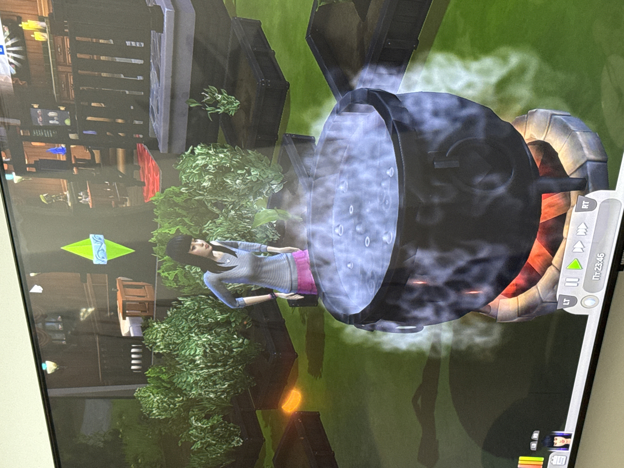 The image depicts a scene from the video game "The Sims," possibly "The Sims 4," displayed on a television screen. A female Sim character, wearing a gray top and pink pants, stands next to a large cauldron. The cauldron appears to be emitting smoke or steam. The Sim is located in an outdoor area with green foliage and part of a house visible in the background. The game's user interface is visible in the corner of the screen.
There is no evidence to suggest that this scene is located in Ramat Gan, Israel, or any specific real-world location.