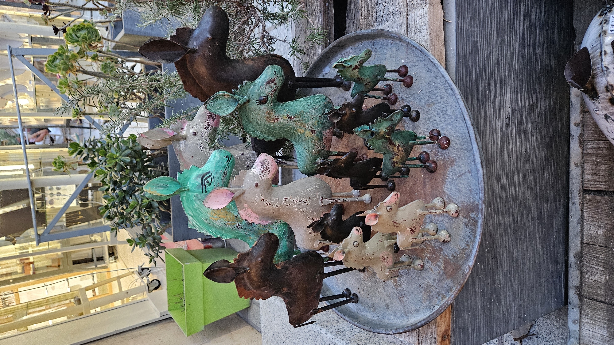 In L'Île-Rousse, France, a collection of handcrafted metal donkey figures are displayed on a round, weathered metal tray placed on a wooden surface. The donkeys vary in size and color, with some painted in metallic green, others in a muted grey with pink accents, and a few in dark brown. They are positioned in a cluster, some facing forward, others turned to the side.
In the background, lush green plants in pots are visible, including succulents and other leafy varieties. Behind the plants, a modern architectural structure with metal beams and glass panels suggests an outdoor retail or exhibition space. The overall scene is outdoors, with natural light illuminating the display. The focus is on the charming, rustic donkey figurines, likely intended for sale as decorative items.