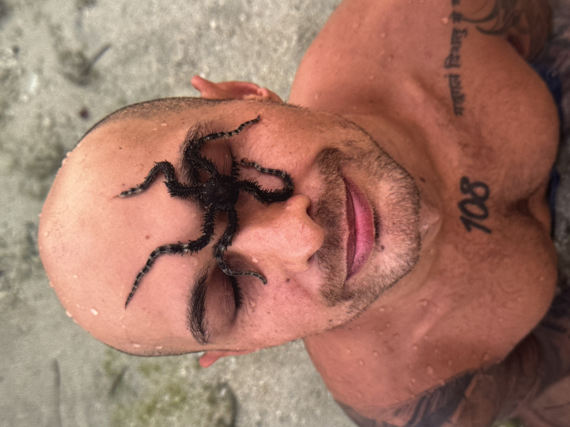 A close-up, top-down shot shows a bald man with a starfish on his face, with its arms extending over his closed eyes and across his forehead. The man is shirtless and appears to be in the water, as evidenced by the water droplets on his skin and the blurred, sandy background that suggests an underwater environment or a shallow beach. He has a tattoo on his chest that reads "108" and another tattoo on his upper chest in an unknown script. The man's expression is neutral, with his lips slightly parted. The image appears to have been taken in Kuta Selatan, Indonesia, possibly during a recreational activity.