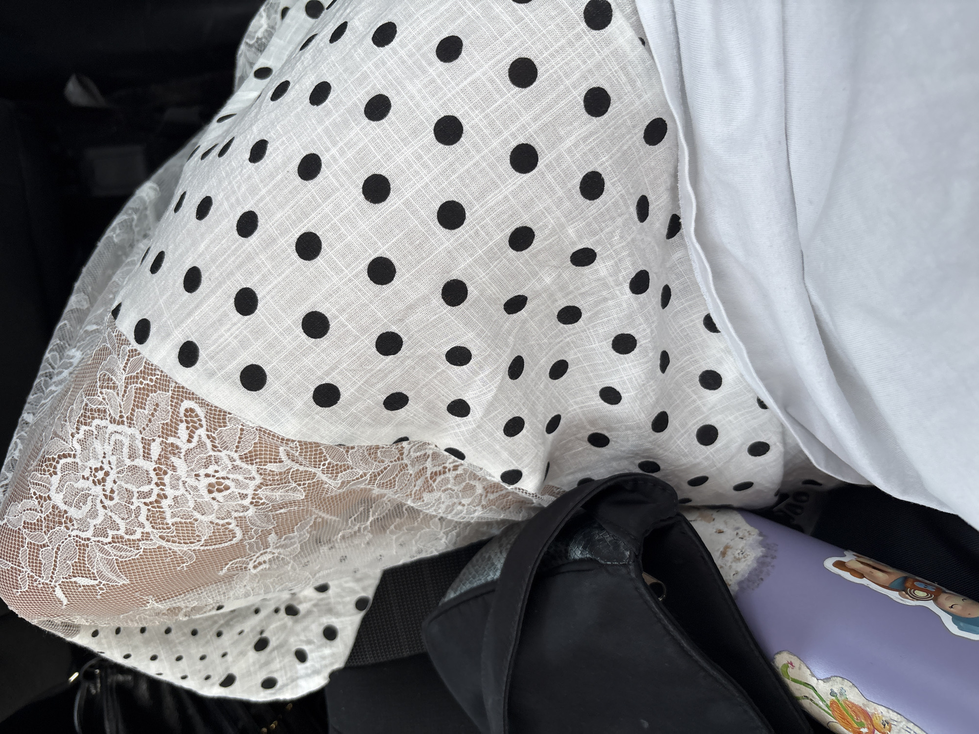 A close-up shot shows a white polka-dot dress with a lace trim overlaying a person's arm. The dress is paired with a white top. A black purse and a lavender-colored object with cartoon stickers are also visible in the lower part of the frame. The location, Huixquilucan de Degollado, Mexico, is not discernible from the image alone.