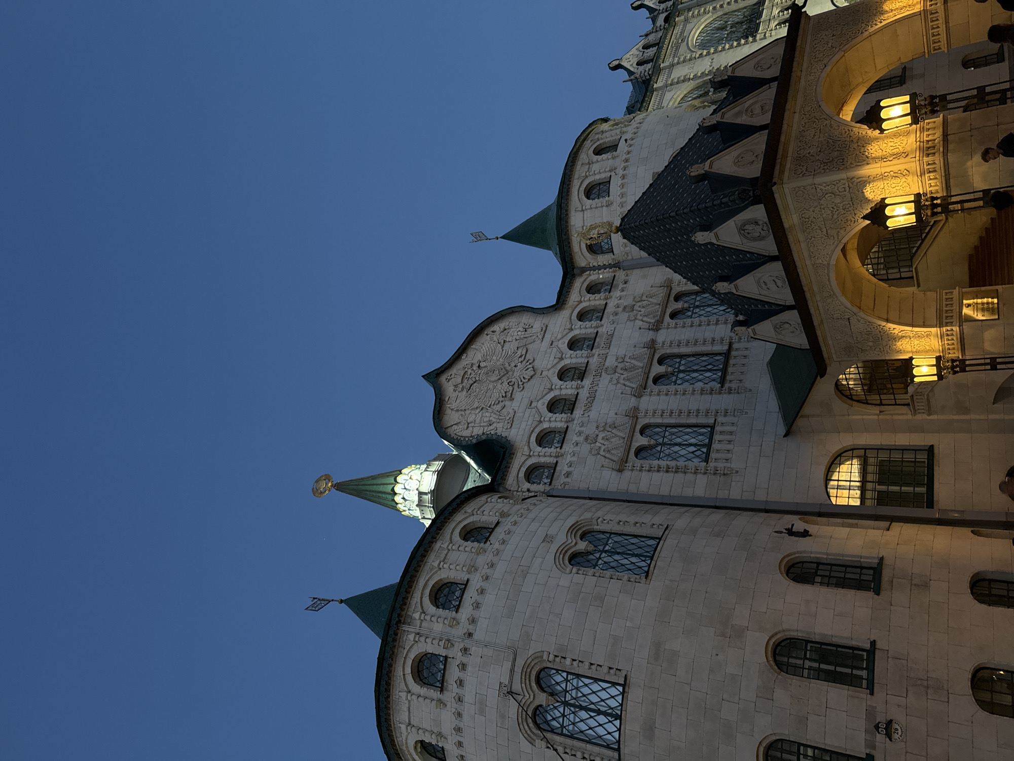 The image shows a low-angle view of an elaborate building against a clear, blue sky. The building features multiple architectural styles, including rounded towers with cone-shaped roofs, arched windows, and ornate facades with relief carvings. Some windows are illuminated from within. The building is constructed from light-colored stone and is likely located in the city of Nizhny Novgorod, Russia, given its distinctive Russian Revival style. Street lights are visible on the right side of the image.