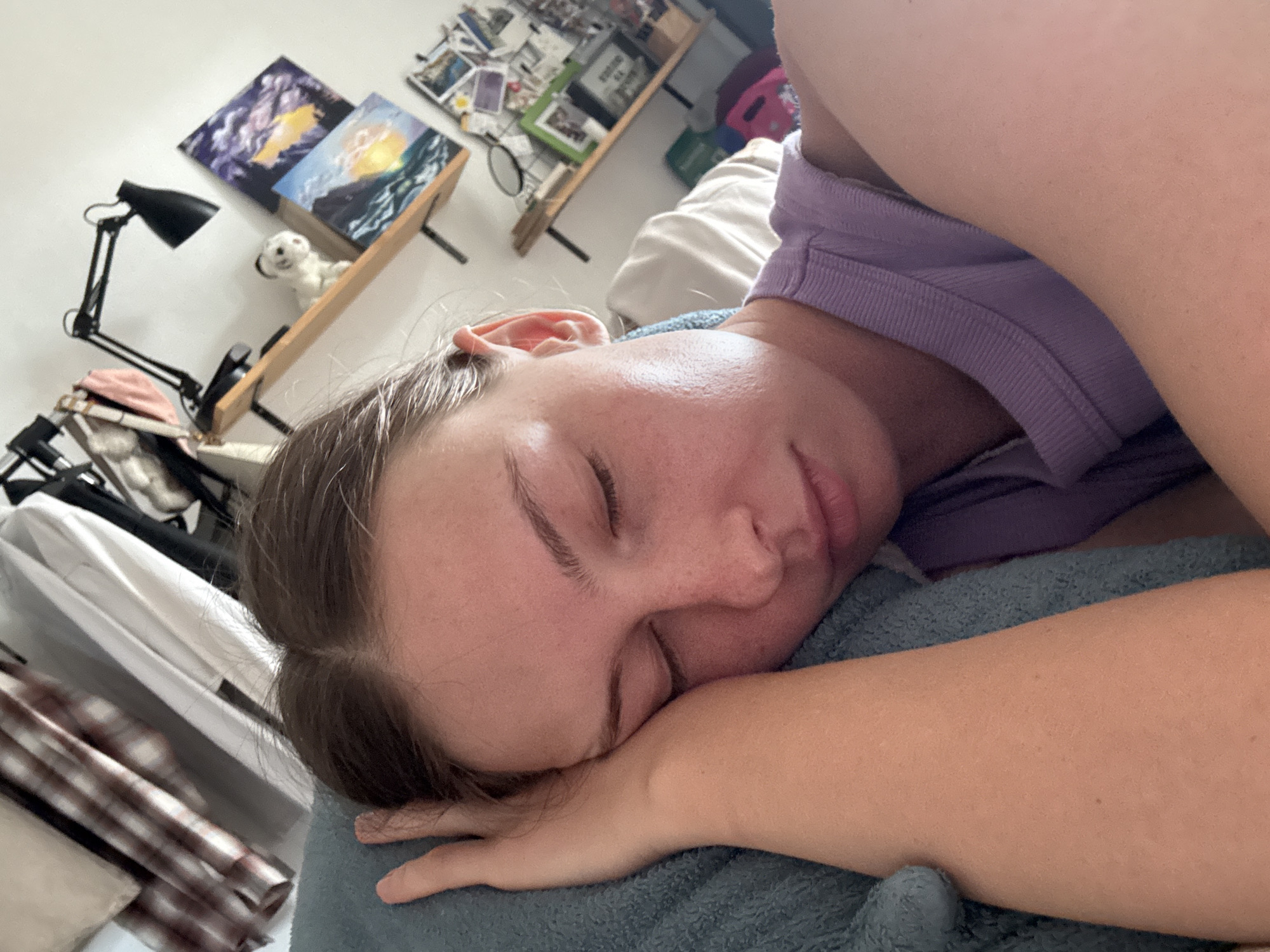 The image captures a close-up perspective of a young adult, appearing female, resting or sleeping on her side. Her eyes are closed, and her head is nestled against a textured, teal-colored pillow or blanket. Her left hand is visible, positioned near her head and resting on the same blue fabric. She has brown hair, parted centrally, and displays light freckles across her nose and cheeks. She is wearing a light purple ribbed sleeveless top.
The background indicates an indoor setting, likely a bedroom or personal room. To the upper left, a clothing rail or rack is visible, from which several garments hang, including a brown and white plaid shirt and a plain white shirt, set against a white wall. To the upper right, a wooden shelf is mounted on a white wall. On this shelf, a black articulated desk lamp is positioned next to a small white decorative figurine, resembling an animal. Two framed or unframed art pieces are displayed on the shelf: the upper one features dark tones with yellow/orange accents, and the lower one depicts a seascape with blue waves and a prominent yellow sun or light source. Below this, another section of the wall or a lower shelf displays various small photographs or postcards, a small circular mirror, and several framed items, one notably with a green frame. A light-colored fabric item is partially visible behind the subject's right arm, and a portion of a pink container is seen in the lower right corner of the background. The lighting is bright and even, suggesting a well-lit interior.