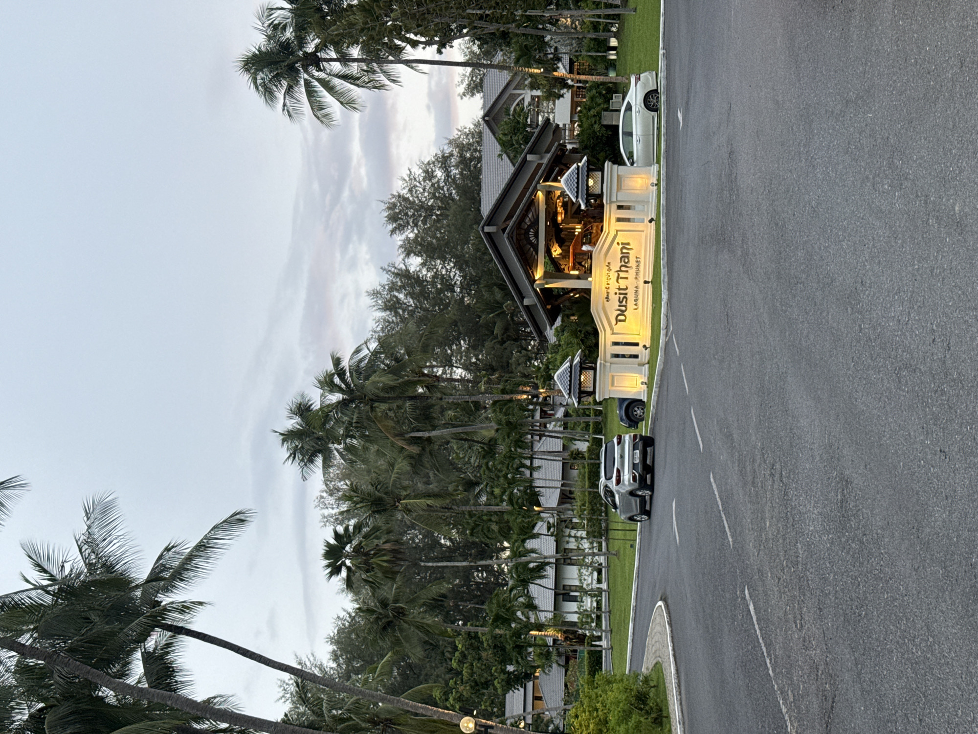 The image shows the entrance to the Dusit Thani Laguna Phuket in Choeng Thale, Thailand, with a road leading to the resort. The sky is overcast with muted colors, suggesting it is late afternoon or early evening. Lush green palm trees and other tropical foliage surround the entrance. A white sign with "Dusit Thani" and "Laguna Phuket" written in gold lettering is prominently displayed at the entrance, illuminated by warm lights. Several cars are visible on the paved road, including a silver SUV and a white sedan. The resort buildings, with their white walls and grey tiled roofs, are visible behind the entrance structure. The scene conveys a sense of arrival at a tropical resort destination.