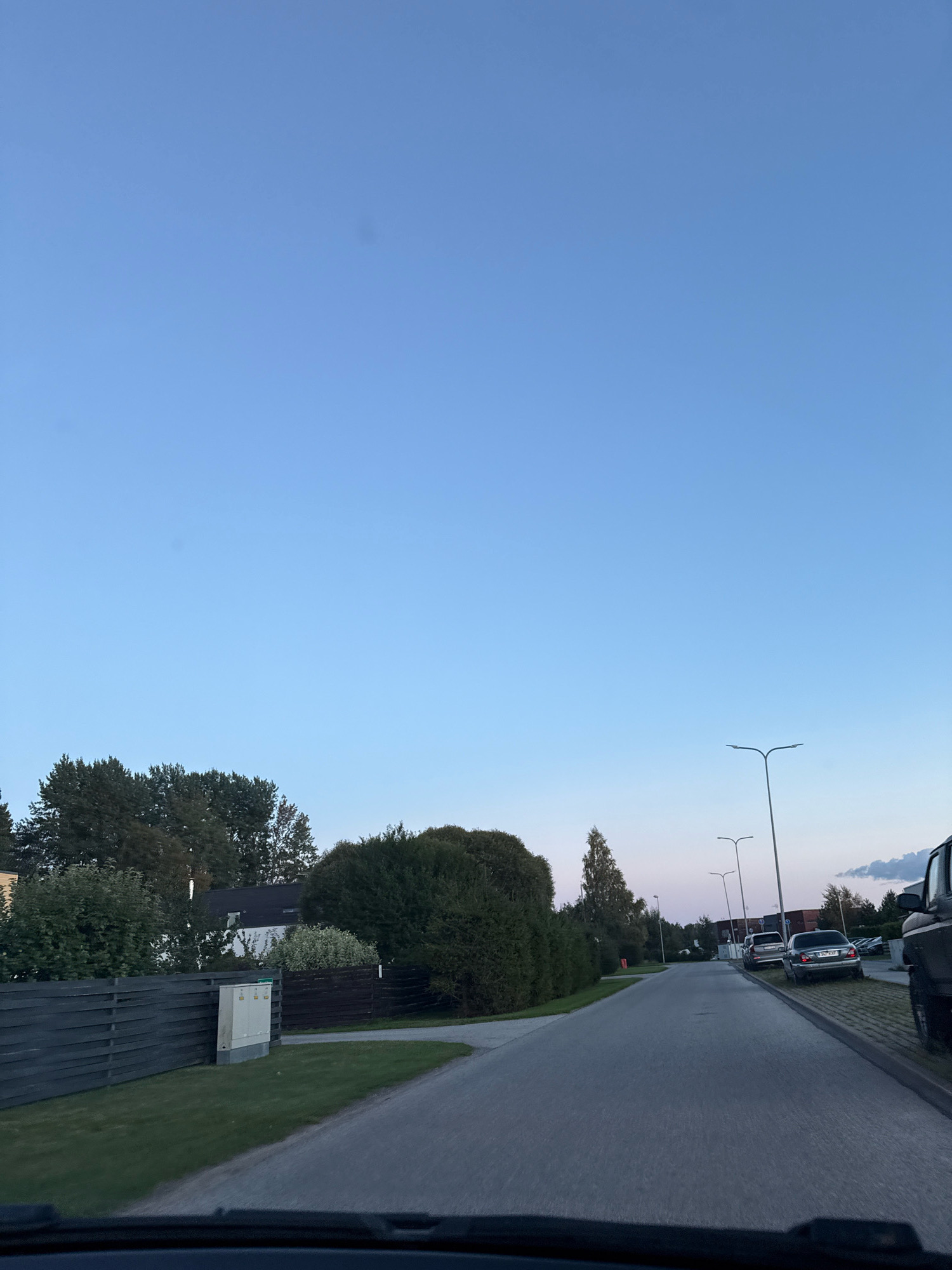 The image depicts a street scene in the city of Alliku, Estonia, likely during twilight. The sky is a clear, light blue with a subtle gradient towards a faint pinkish hue near the horizon. On the left side of the road, a dark wooden fence runs along a grassy area, with a white utility box situated near it. Beyond the fence, lush green trees and some houses are visible. The road itself is paved and curves gently to the right, with several parked cars lining the right side. These vehicles include a dark-colored SUV, a silver sedan, and a few other cars further down the street. Streetlights with a modern design are positioned along the road. The overall impression is a quiet residential or suburban area.