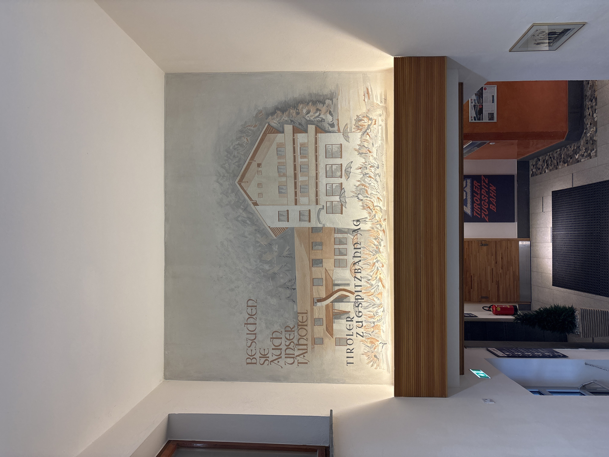 The image shows an interior view of a building, likely a hotel or a public establishment, with a large mural painted on the wall above a wooden beam. The mural depicts a snow-covered alpine scene with a hotel building, trees, and text that reads "Besuchen Sie auch unser Talhotel" and "Tiroler Zugspitzbahn AG". Below the mural, a wooden beam separates the artwork from the lower part of the building, which features a staircase and a hallway with an exit sign, a fire extinguisher, a potted plant, and a framed picture on the wall. The overall impression is one of a welcoming and rustic mountain-themed environment. Based on the text on the mural, this location is likely in Ehrwald, Austria, given its connection to the Tiroler Zugspitzbahn, a cable car railway that goes up the Zugspitze mountain.