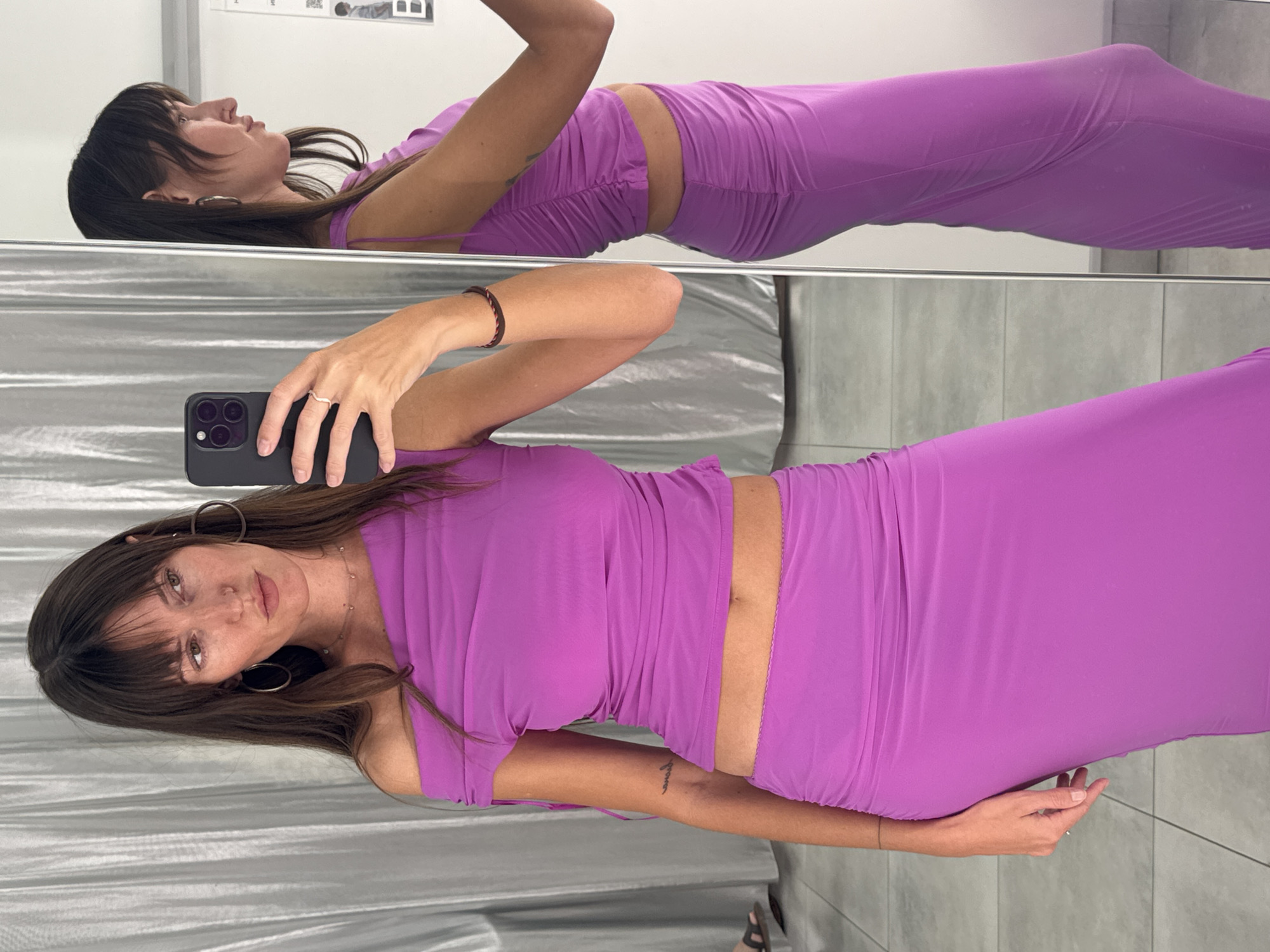 A woman with long brown hair and bangs is taking a selfie in a store. She is wearing a bright purple off-the-shoulder top with ruching and a matching ruched maxi skirt. She has hoop earrings, a delicate necklace, and rings on her fingers. The background appears to be a fitting room with a silver curtain and a full-length mirror. The woman is standing in front of the mirror, and her reflection is visible to the right of the image. The floor is tiled. The overall scene suggests a shopping environment. The location is identified as Khimki, Russia.
