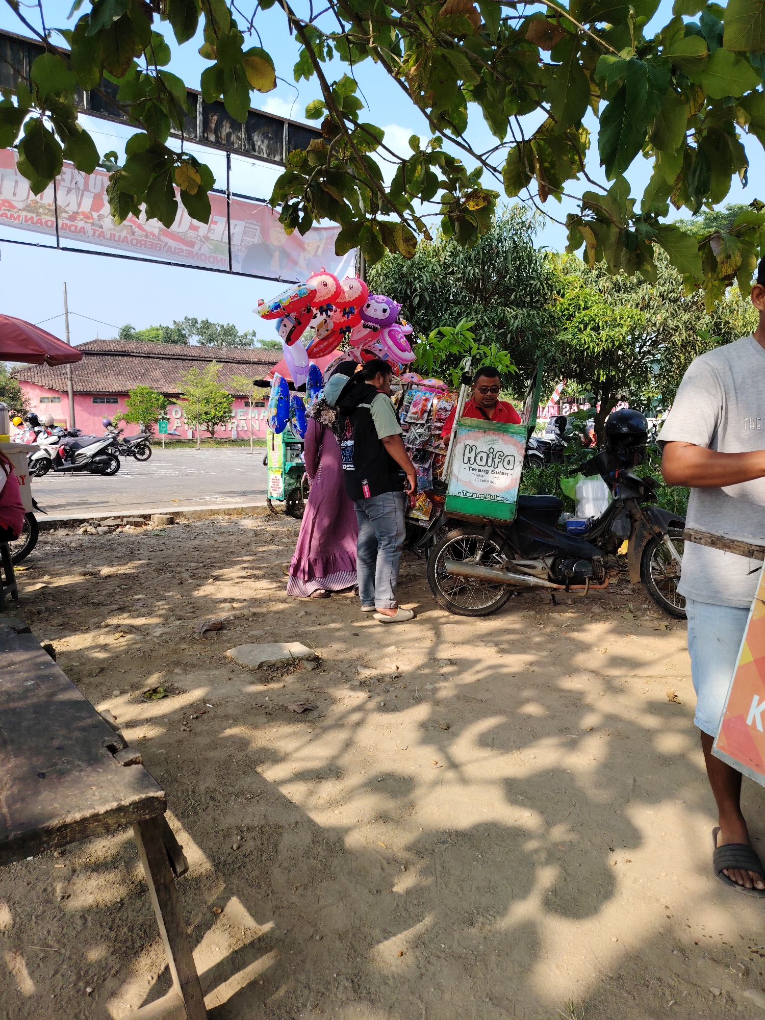 In Semarang, Indonesia, under the shade of trees, a street scene unfolds. A vendor pushes a cart laden with colorful balloons, a mix of character-themed and round designs, along a dirt path. Beside the cart, a man in a red shirt and sunglasses oversees the operation, while another individual in a purple dress stands near the balloons. A motorbike with a large box on the back is parked next to the cart, displaying the brand "Haifa Terang Bulan". In the background, a pink building with a tiled roof is visible, with parked motorcycles in front of it. The ground is a mix of dirt and gravel, with shadows cast by overhead foliage. A portion of a wooden table and a person in a grey t-shirt and blue shorts are also in the foreground.