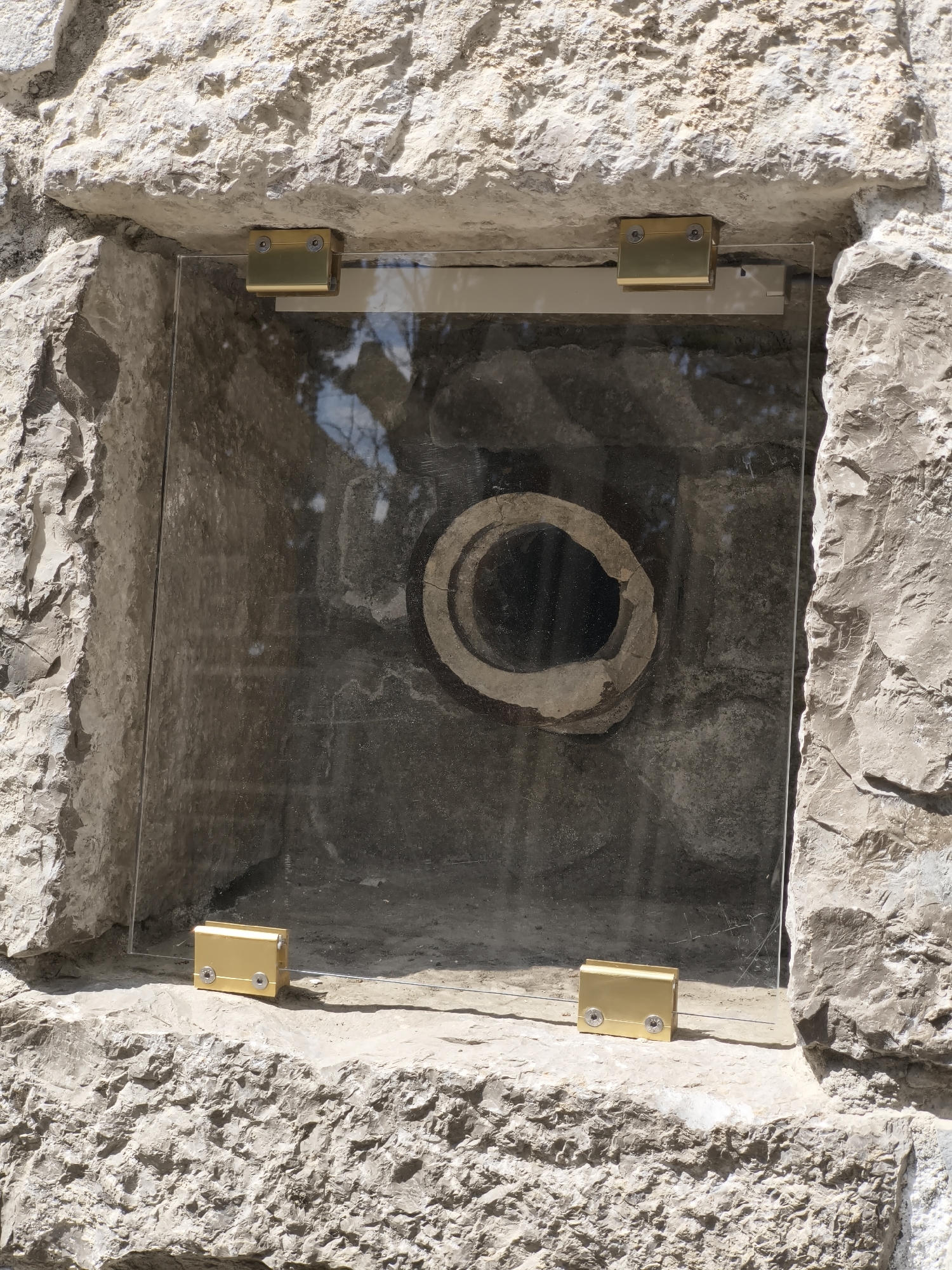 A rectangular alcove in a light-colored stone wall holds a transparent, possibly glass, panel secured by four gold-colored metal brackets. The panel obscures, yet partially reveals, the dark interior of the alcove, featuring a circular stone or clay structure with a black hole at its center. The surrounding wall appears rough and weathered, suggesting an outdoor location. The location is in the city of Unknown, Unknown.