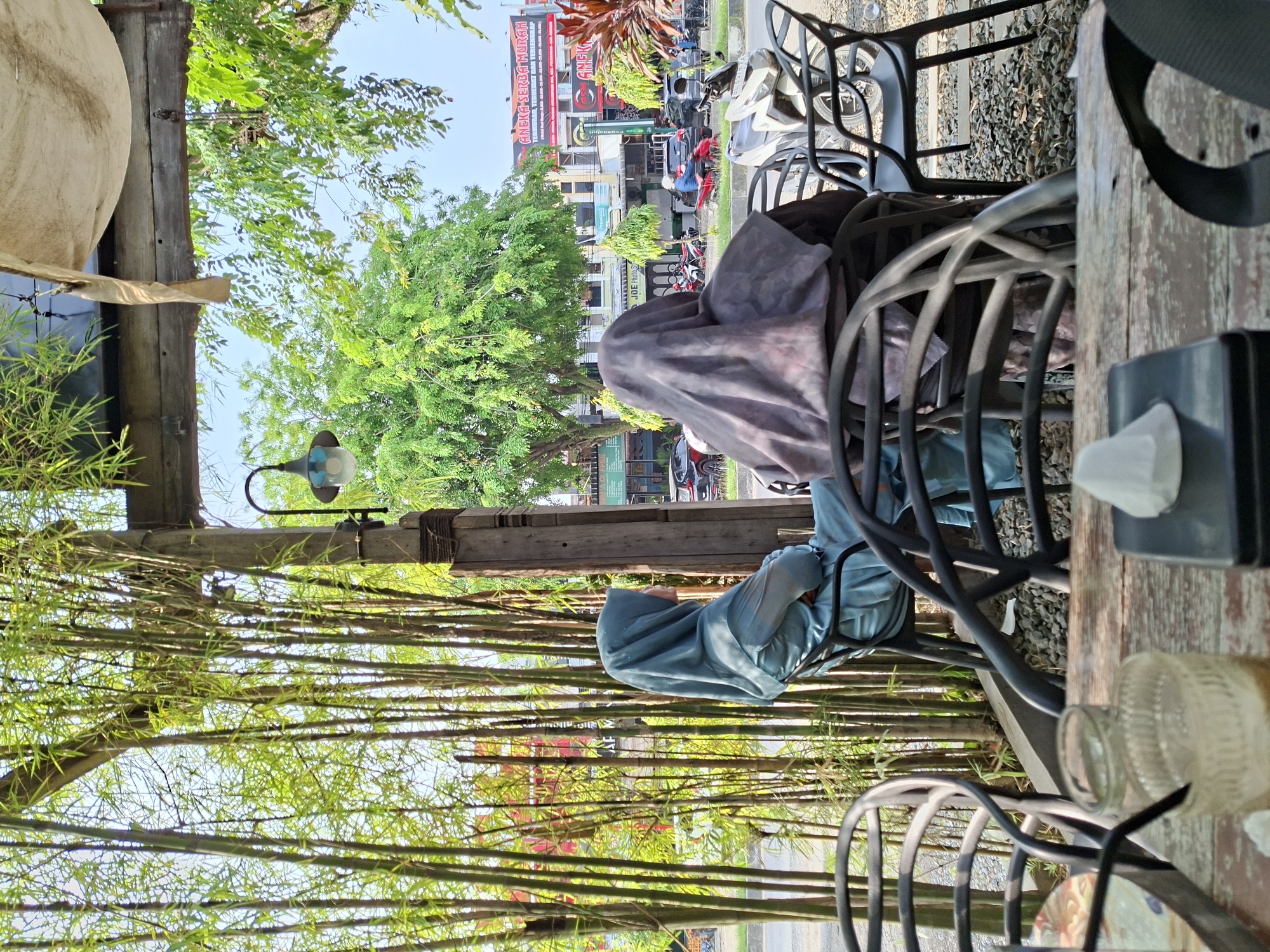 The image shows an outdoor cafe in Pantegajah, Indonesia. A person wearing a blue hijab is seated at a wooden table with black metal chairs. Another chair is covered with a gray cloth. A menu and a condiment container are on the table. The cafe is decorated with bamboo stalks. Across the street, buildings are visible. Motorbikes and cars are passing by.