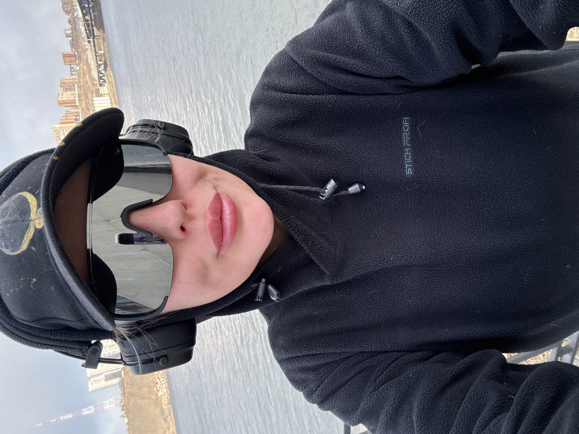 A person is captured in a close-up selfie, oriented with their head tilted slightly to the right, against an outdoor background. They are wearing a black fleece hoodie or jacket, a black baseball cap with a subtle gold-toned design on the side, large reflective mirrored sunglasses, and black over-ear headphones. The sunglasses obscure their eyes, but their nose and full lips with a soft pink tint are visible.
The background consists of a wide body of water, likely a river, reflecting a soft, diffuse light that suggests an overcast day. On the distant bank, a line of buildings and industrial structures can be seen, hinting at an urban or semi-urban environment. This landscape is consistent with the Yenisei River in Krasnoyarsk, Russia.
The black fleece jacket features a drawstring at the neck with two black toggles. On the left chest area, the brand name "STICH PROFI" is subtly embroidered in dark thread. The overall impression is one of casual comfort in cool weather, perhaps while engaging in outdoor activity like walking or commuting.