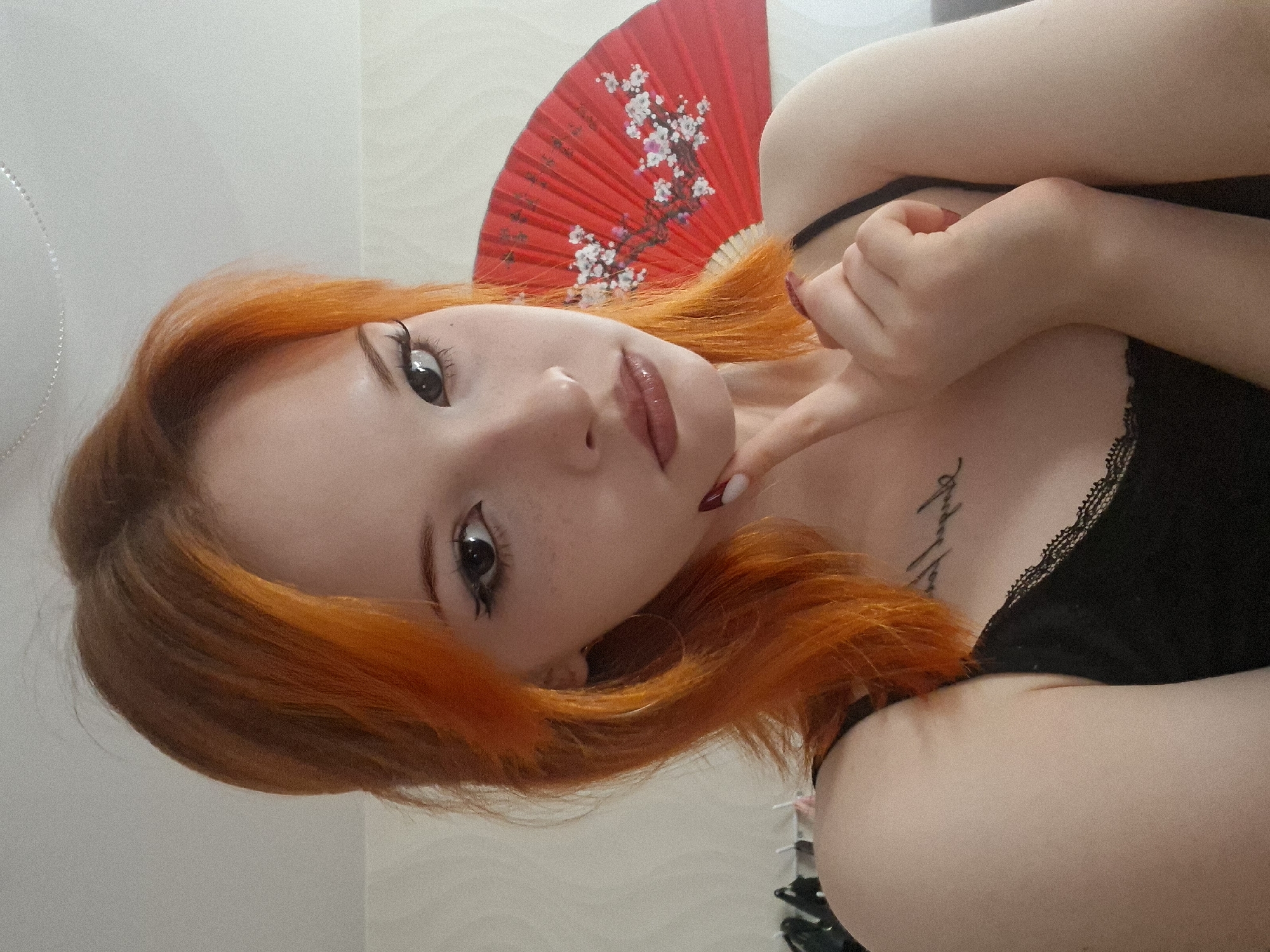 A young woman with bright red hair is holding a red and white Asian-style fan behind her head. She has fair skin and is wearing a black top with lace trim. She has dark eyeliner and glossy lips. Her left hand is raised near her face, with her index finger pointing towards her chin. There is a tattoo visible on her chest. The background is a plain white wall. The location is not identifiable as the city of Chaikovsky, Russia.