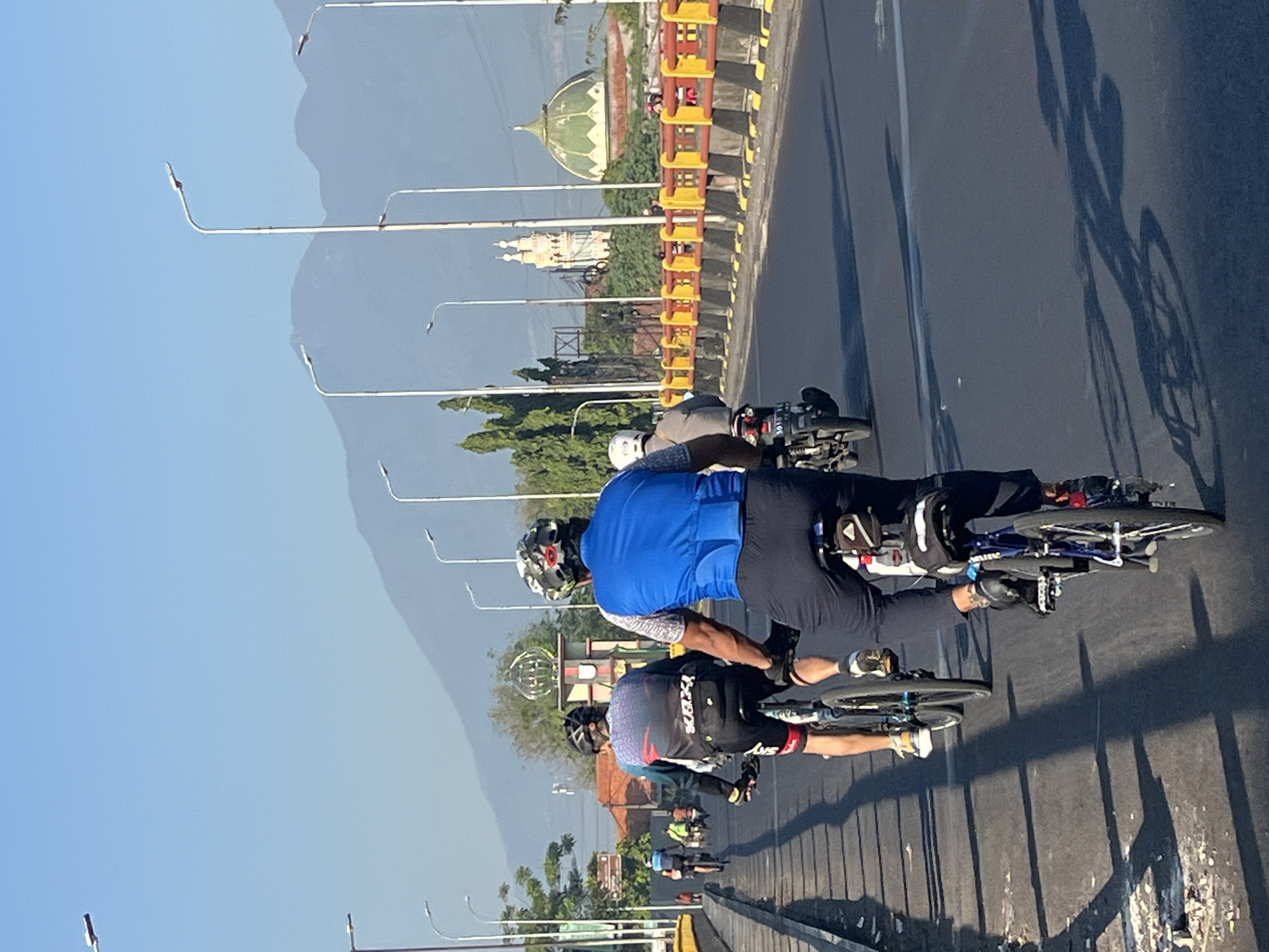 This is a photograph taken in Gempol, Indonesia, capturing a group of cyclists on a bridge. The perspective is from behind a cyclist in the foreground, who is wearing a blue vest and a helmet. The cyclist is on a bicycle with a blue frame and is facing away from the camera, riding on the right side of the road. To the left of the foreground cyclist is another cyclist wearing a patterned jersey and black cycling shorts. Behind them, several other cyclists and a motorcycle are visible on the bridge, moving in the same direction. The bridge has a yellow and red guardrail along its side, and streetlights line the roadway. In the background, lush green trees and buildings, including a structure with a green dome and a tall tower, are visible. The sky is clear and blue, and mountains are faintly discernible in the distance. The sun casts long shadows of the cyclists and their bicycles onto the asphalt road.