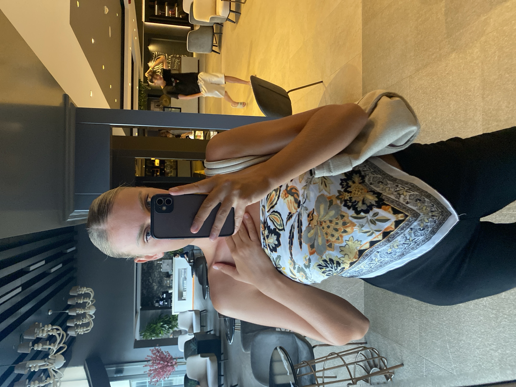 A young woman with blonde hair pulled back poses for a mirror selfie in a modern, well-lit lobby area. She is wearing a strapless scarf top with a colorful floral pattern and black leggings. Her right arm is raised, holding a black smartphone in front of her face, and her left hand is placed on her chest. A beige shoulder bag is slung over her left shoulder. In the background, reflected in the mirror, a young man in a black t-shirt and white shorts is walking, and other seating areas and decor are visible. The floor is tiled in a warm, yellow tone, and the ceiling features dark beams and multiple light fixtures. The woman is positioned in front of a dark, reflective column. The overall impression is of a hotel or commercial space with contemporary design elements. The location is the lobby of the AKSU, Turkey city hotel.