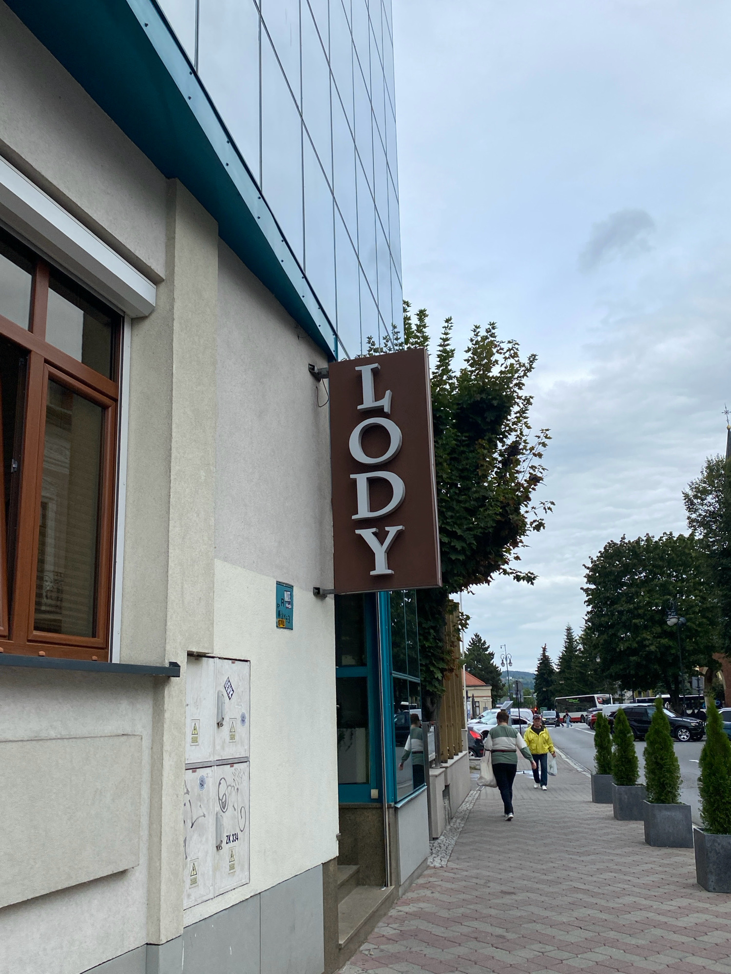 This is a street-level, eye-level, medium shot of a building facade on a cloudy day in Nowy Sącz, Poland. The building has a modern glass facade on the upper levels and a textured beige and grey lower facade. A brown sign with white lettering that reads "LODY" (Polish for "ice cream") is mounted on the building. To the left of the sign is a window with brown trim, and below that is an electrical utility box with warning stickers and graffiti. A sidewalk with red and grey brick paving runs in front of the building, lined with planters containing conical green trees. Two people are walking on the sidewalk away from the camera. The person closer to the camera is wearing a striped sweater and dark pants, carrying a white bag. The person further away is wearing a yellow jacket and dark pants, also carrying a white bag. In the background, there is a street with traffic, including cars and a bus, and more trees and buildings.