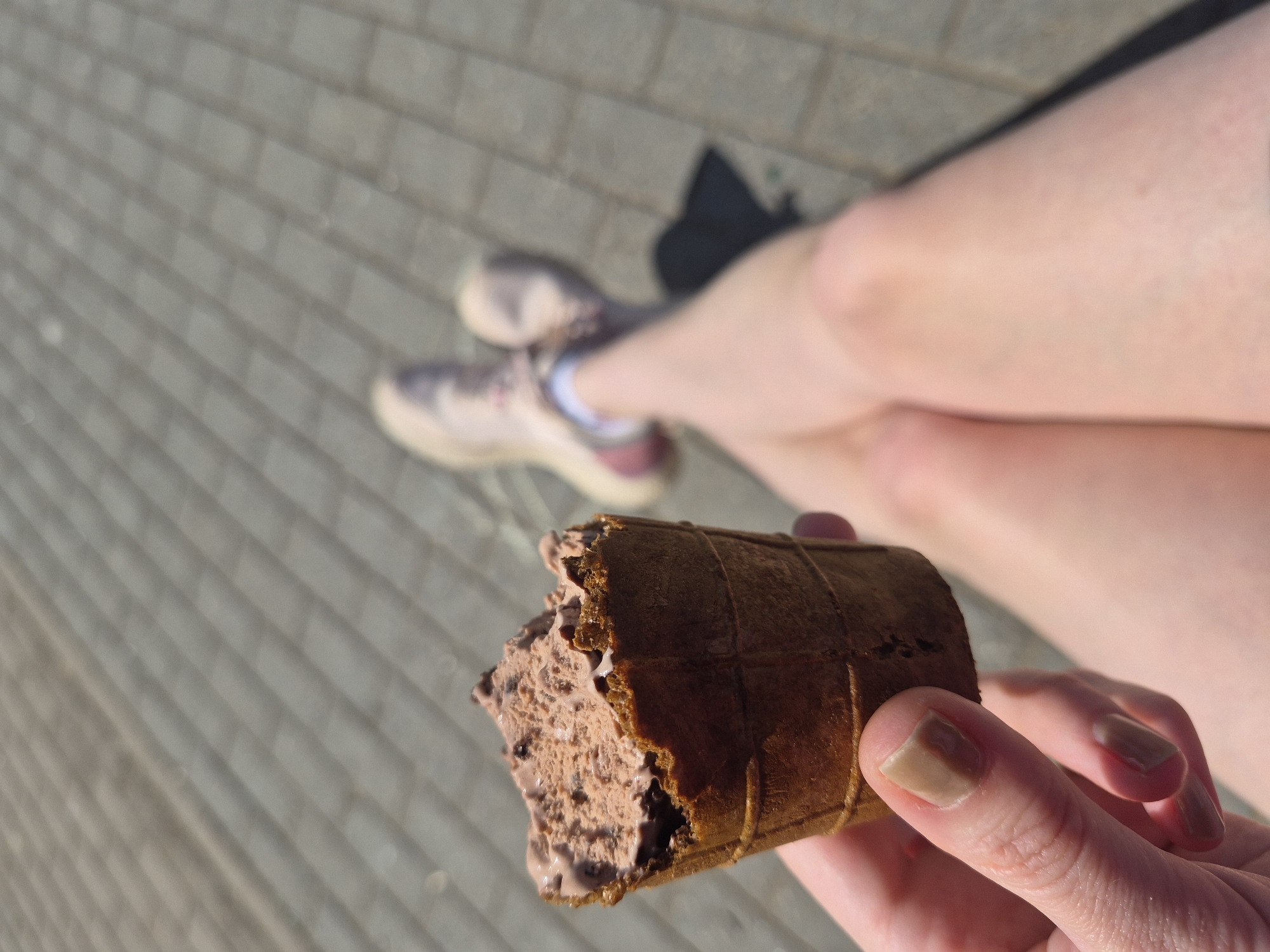 A person is holding a chocolate ice cream cone with a bite taken out of it. The background shows a person's legs, feet in sneakers, and a paved surface with brick-like patterns. The location is an outdoor space, possibly a street or plaza in an urban environment.