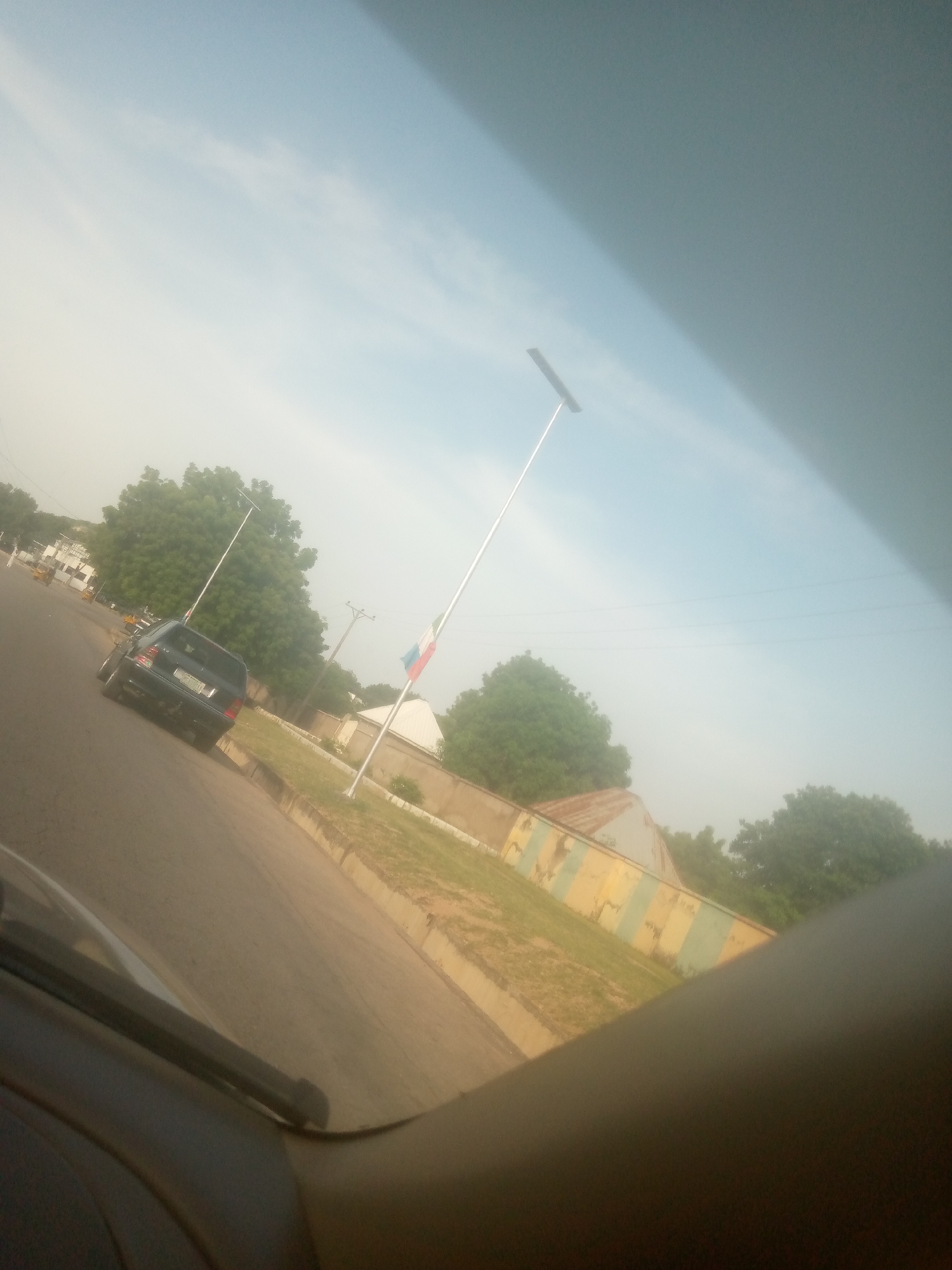 The image depicts a street scene, likely in Jalingo, Nigeria. A dark-colored SUV is the primary subject, driving on a paved road. The vehicle appears to be a 2007-2014 Mercedes-Benz M-Class. In the background, several large trees are visible, suggesting a somewhat rural or suburban setting. A tall flagpole with a partially visible flag, which appears to be red, white, and blue, stands to the right of the road. The sky is a pale blue with some wispy clouds, indicating it is daytime, likely late morning or early afternoon given the lighting. There's a building with a distinctive striped facade (yellow and green) behind the flagpole. The image is taken from within another vehicle, as part of a car interior is visible in the foreground. The overall atmosphere seems calm and ordinary, with no specific event or activity being clearly depicted beyond a vehicle in motion.