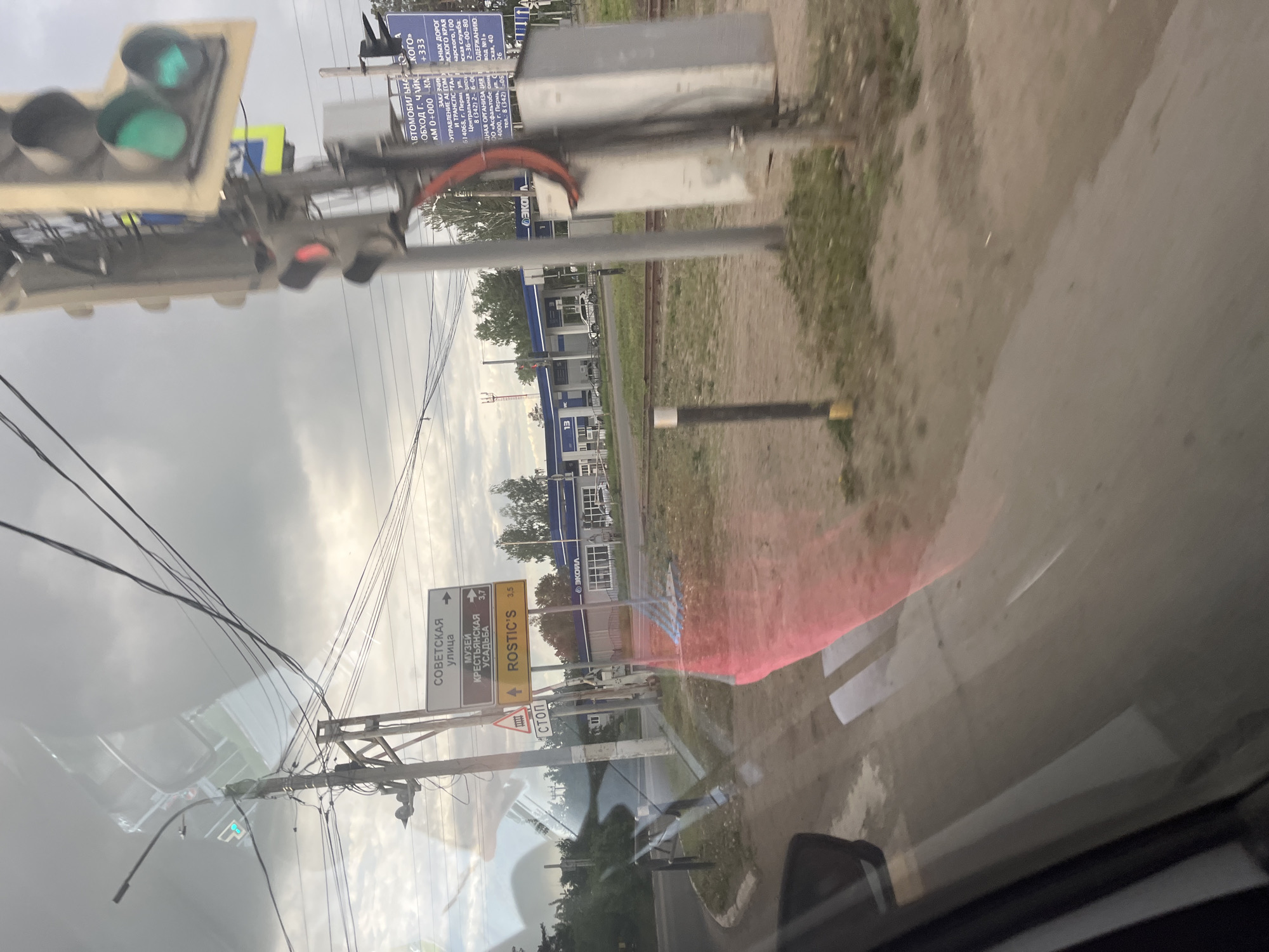 The image, taken from inside a vehicle, shows a street scene in Chaikovsky, Russia. A traffic light displays green. Street signs indicate "SOVETSKAYA" street, "MUSEUM OF THE PEASANT ESTATE," and "ROSTIC'S." Another sign provides information related to the road "АВТОМОБИЛЬ (AUTOMOBILE) ЧАЙКОВСКОГО," with the kilometer marker "KM 0+000 KM." Further text describes "Обход г. Чайковского (Chaikovsky Bypass)." There is a pedestrian crosswalk. A blue gas station is visible in the background. Power lines crisscross the sky.