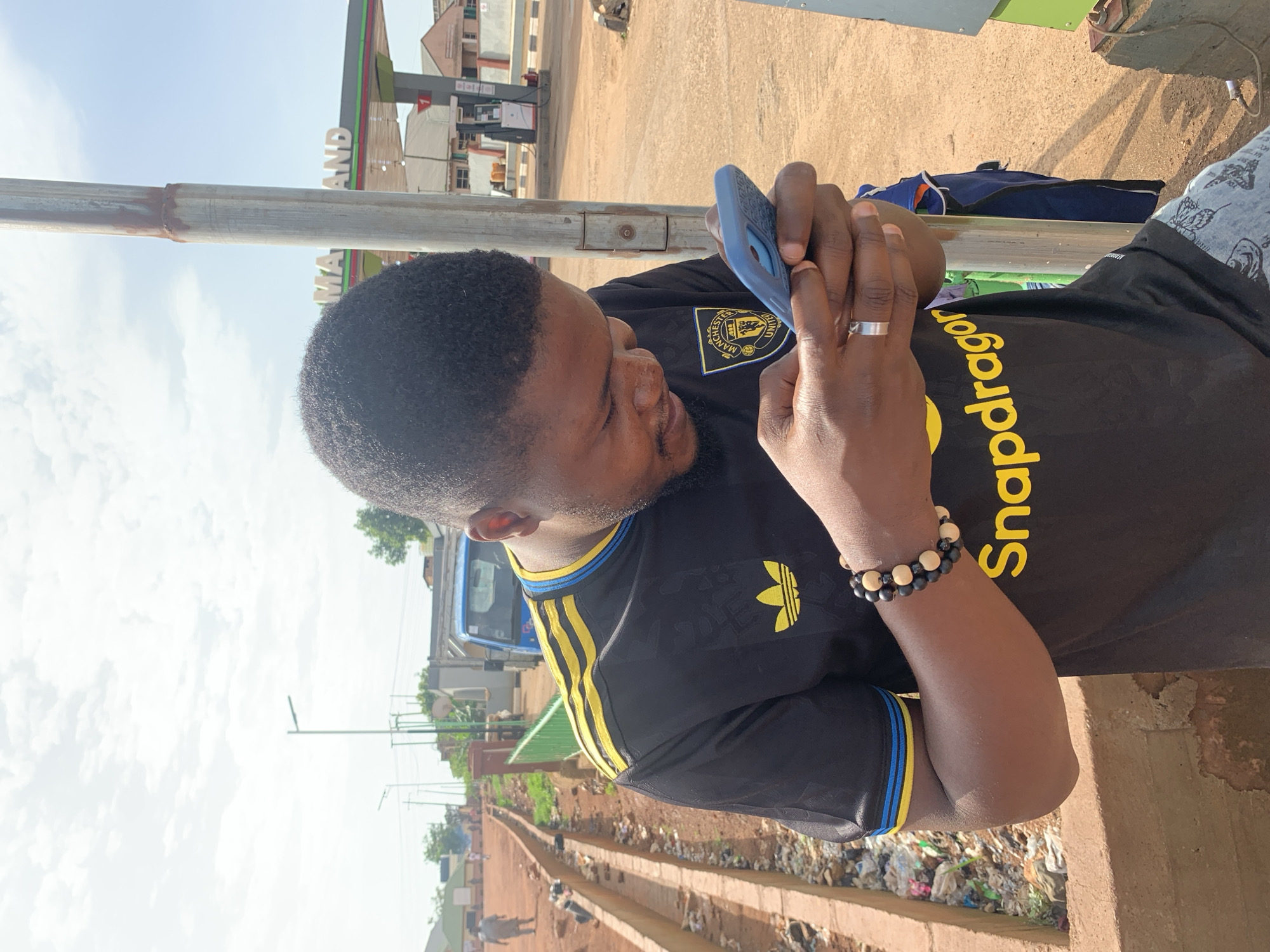 A young Black male is pictured from the chest up, outdoors, looking down at his blue smartphone. He is wearing a black t-shirt with yellow and blue accents on the collar and sleeves. The t-shirt also features a yellow Adidas logo and "Snapdragon" text in yellow lettering. He has on a beaded bracelet on his left wrist and a silver ring on his left ring finger.
The background shows a street scene in Gura Top, Nigeria. To the left, a rough embankment lines a drainage ditch filled with refuse. Further down the road, a blue truck is parked, and other vehicles are visible. A gas station with a "MAZAND" sign is in the background on the right. The sky is cloudy. The man appears to be standing next to a metal pole.