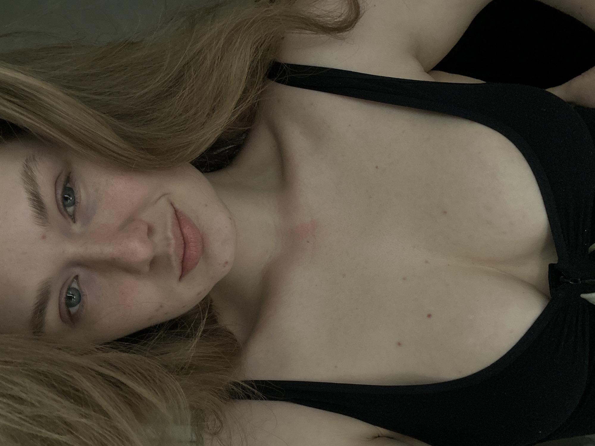 A close-up, slightly angled, eye-level shot presents a woman with fair skin and light brown hair, lying down. She is wearing a black top with a low neckline. Her face is tilted towards the camera, featuring light blue eyes, freckles, and a gentle smile. The background is dark and out of focus. There is no indication of Kazan, Russia in the image.
