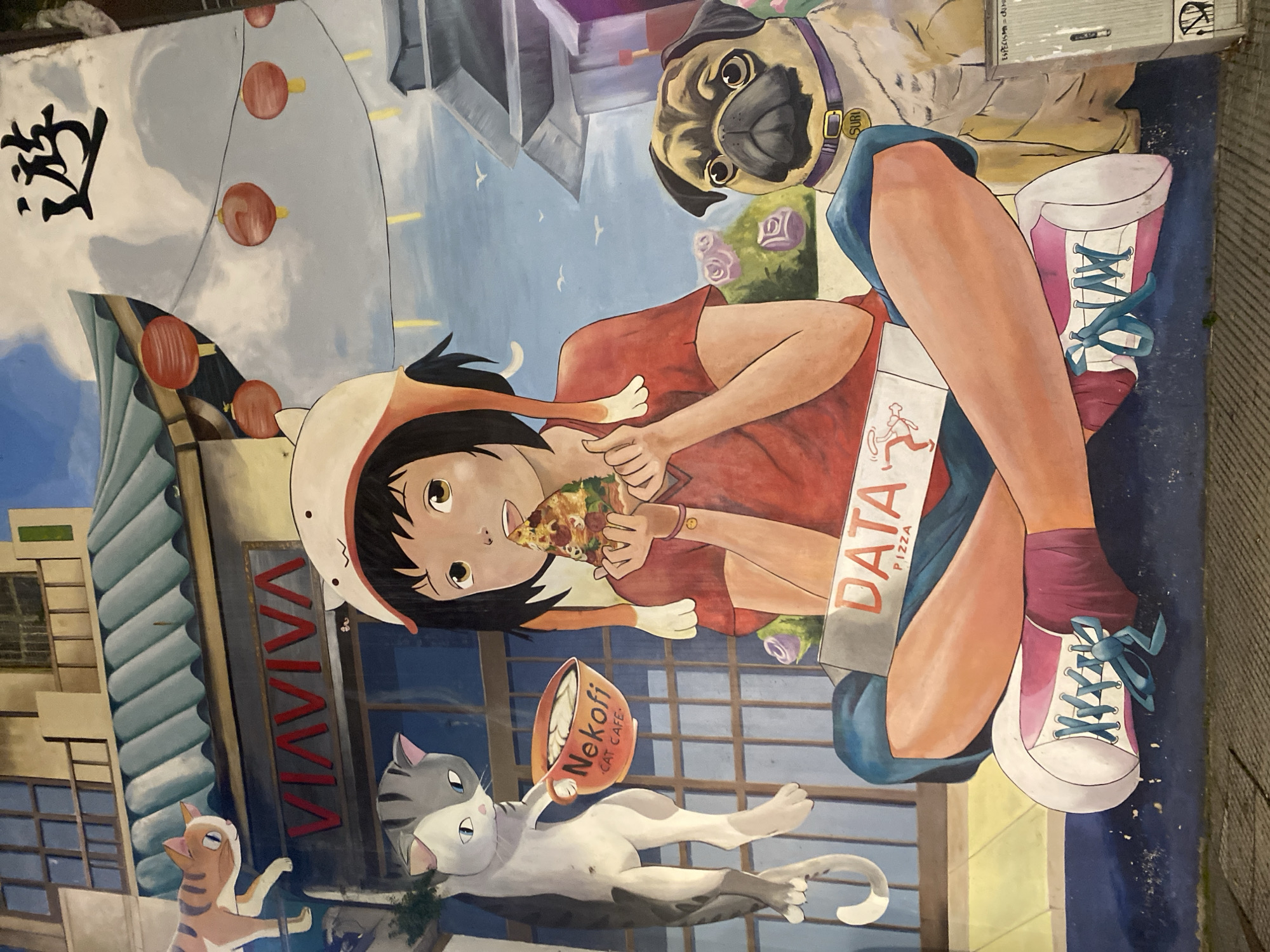 This is a street art mural in Buenos Aires, Argentina. The mural depicts an anime-style girl in a cat-eared hat eating pizza. Next to her is a pug wearing a collar with "SURI" on it, and a box labeled "DATA PIZZA" beside her legs. Below her, a white cat lies next to an orange cup labeled "Nekofi CAT CAFE." Another cat appears to be climbing the side of a building with the sign "VIVIVA" in red letters. Lanterns hang near the building. Some buildings are visible in the background along with some birds in the sky. A black character is written above the building.