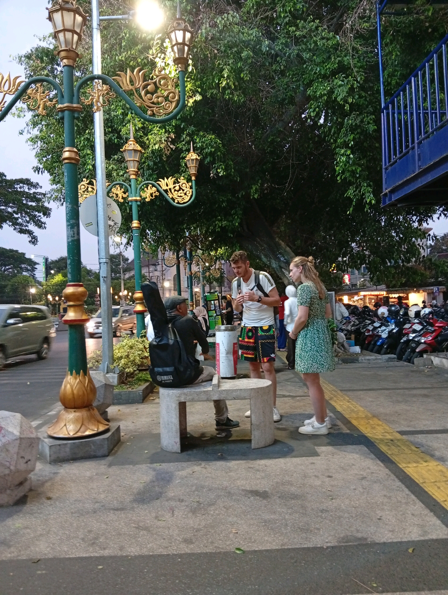 The image captures a street scene in Malang, Indonesia, likely in the evening. In the foreground, a man wearing a flat cap and a black long-sleeved shirt is seated on a stone bench, with a black guitar case with a "YAMAHA" logo propped behind him. He appears to be looking to his right. Two other individuals, a man and a woman, are standing and interacting. The man, with a beard, wears a white t-shirt and patterned shorts. The woman is in a green patterned dress. Behind them, a row of parked motorcycles is visible, and further back, a blue elevated structure with railings looms. The street is lit by ornate lampposts, and trees with dense foliage line the sidewalk. Cars are seen driving on the road in the background, indicating a public area. The overall atmosphere suggests a lively urban environment.