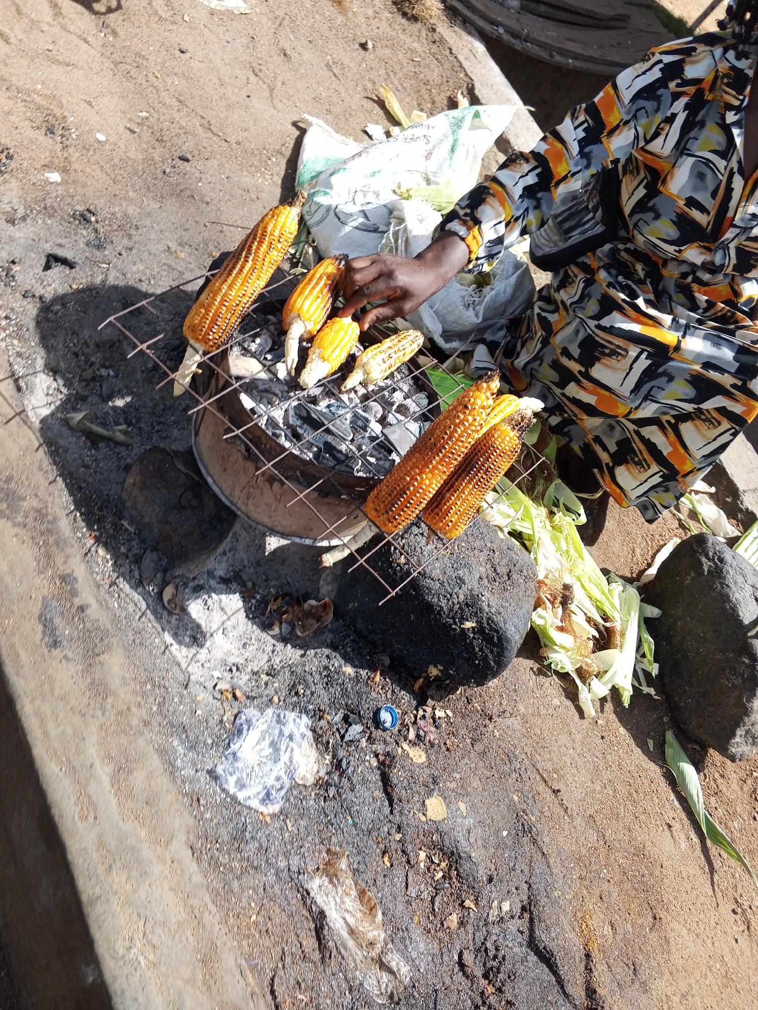In Bukuru, Nigeria, a woman wearing a patterned, long-sleeved dress is grilling corn on a charcoal grill. The grill, supported by stones, holds four partially grilled corn cobs. Several more cobs and corn husks are visible near the grill. The surrounding area is a mix of concrete and dirt, with scattered debris and a piece of plastic wrap.