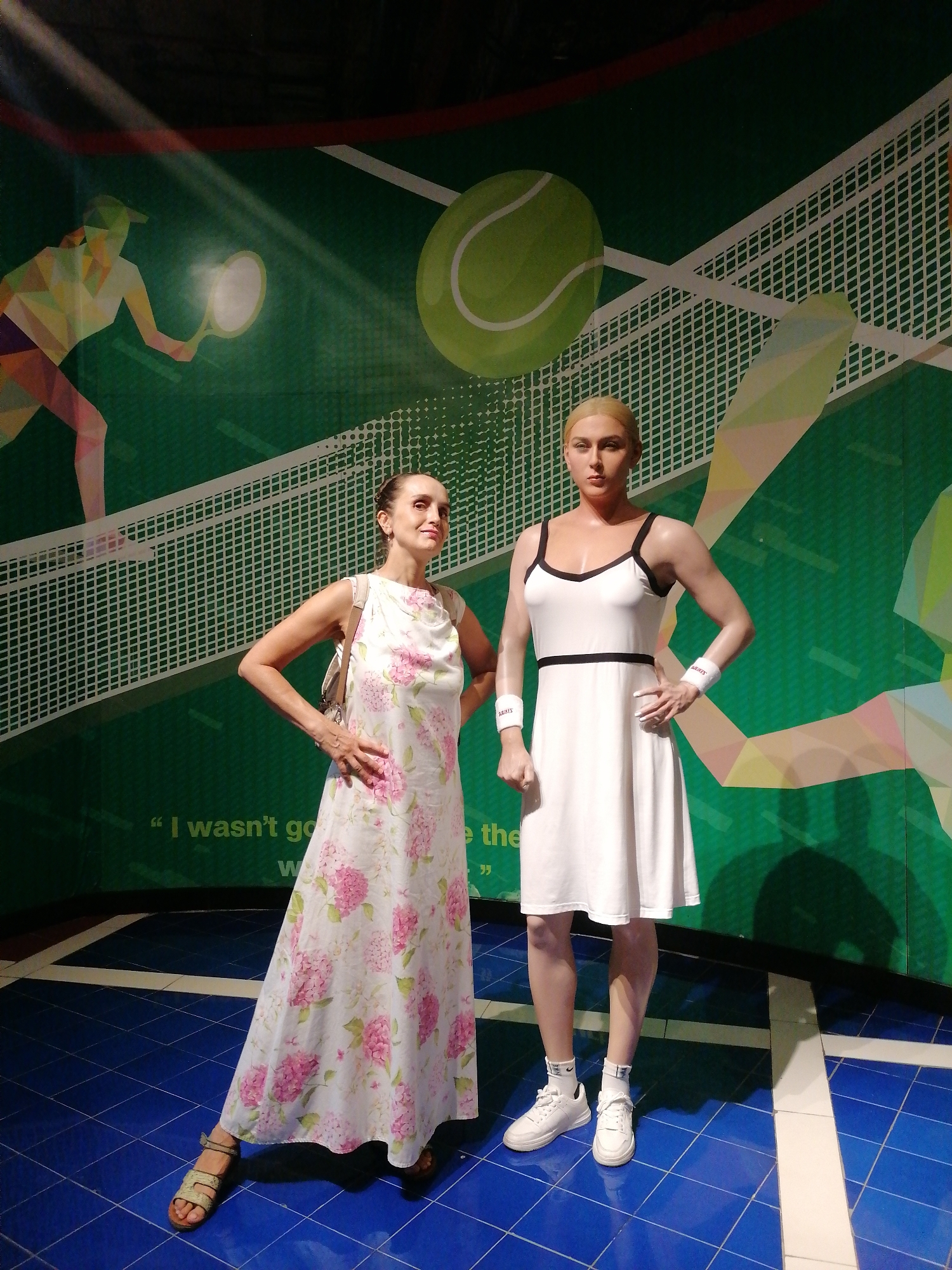 A woman stands next to a wax figure of a tennis player. They are positioned in front of a backdrop depicting a tennis court with a net, a tennis ball, and a figure of a tennis player. The location is the wax museum in an Unknown city.
The woman on the left is wearing a long white dress with a floral pattern and has light brown sandals on her feet. Her hair is pulled back, and she has her hands on her hips, posing for the picture. The wax figure of the tennis player on the right is wearing a white tennis dress with black trim, white socks, and white sneakers. The wax figure is a likeness of Maria Sharapova. Her hair is blonde, and she has wristbands on both wrists with the word "SHARAPOVA" printed on them. The backdrop is predominantly green, with white lines indicating the net and other tennis court markings. A quote is visible on the backdrop, partially obscured by the woman in the floral dress. The floor has a blue and white checkered pattern.