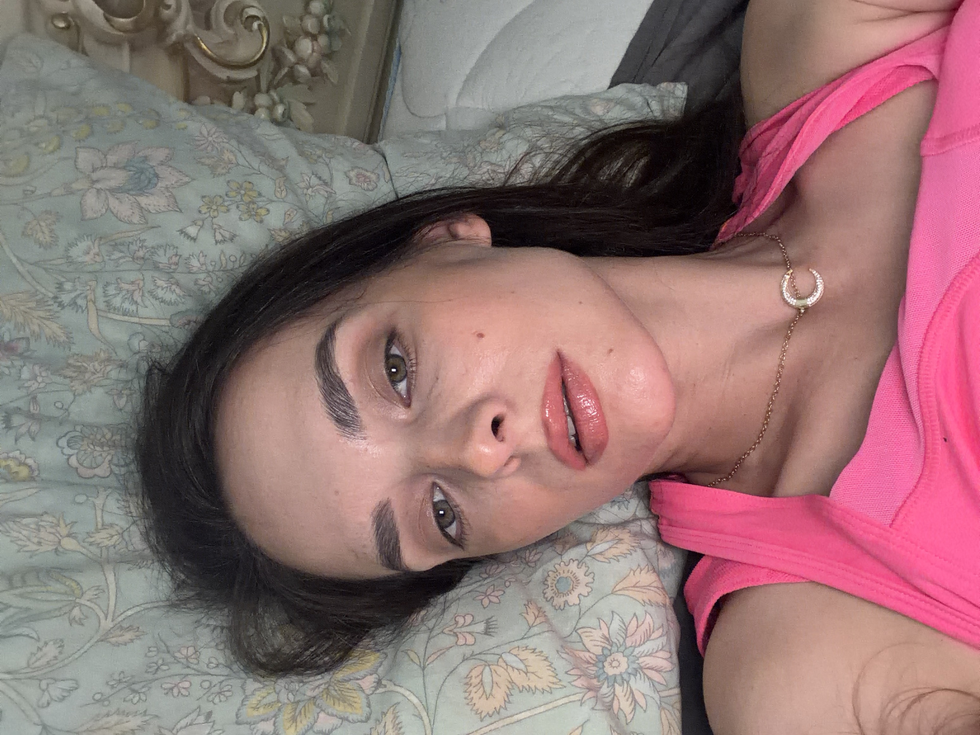 A close-up, overhead, selfie-style shot features a young adult woman with dark hair and light skin lying on a bed. She is wearing a bright pink tank top and a gold necklace with a crescent moon pendant. Her makeup includes neutral eyeshadow, mascara, and a glossy pink lipstick. The background consists of a patterned light green pillow and a portion of a white upholstered headboard. The lighting is soft and appears to be natural, illuminating the woman's face from above.
