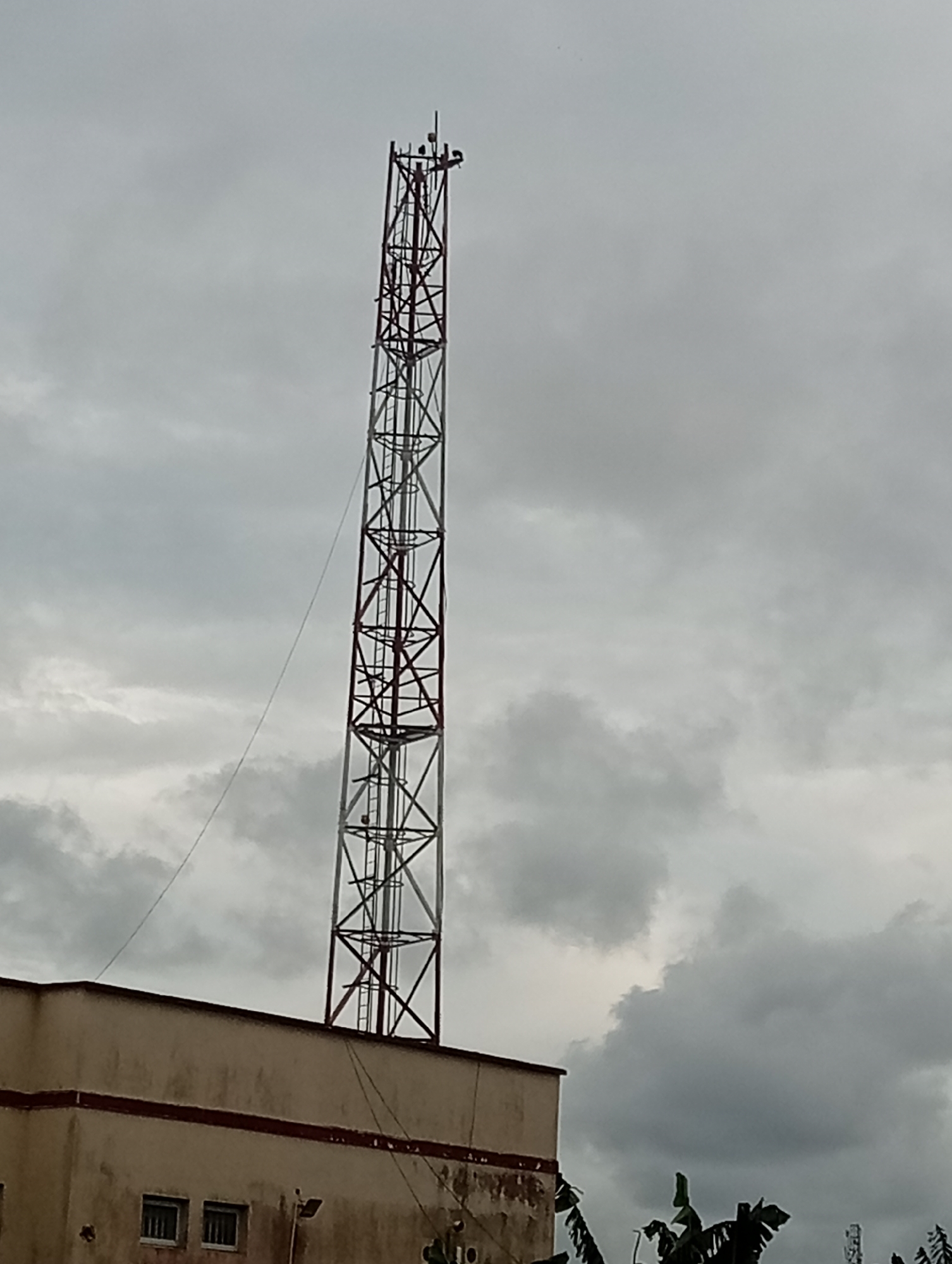 The image depicts a tall telecommunications tower extending high into a cloudy sky. The tower is constructed of a lattice framework, alternating sections of white and red coloring. A ladder runs vertically along one side. The base of the tower appears to be situated on the roof of a two-story, beige-colored building with a horizontal, red accent line. The building has windows and a light fixture are visible. The location is identified as Ijanikin, Nigeria. The lower right corner of the frame reveals foliage, suggesting a somewhat lush environment.
