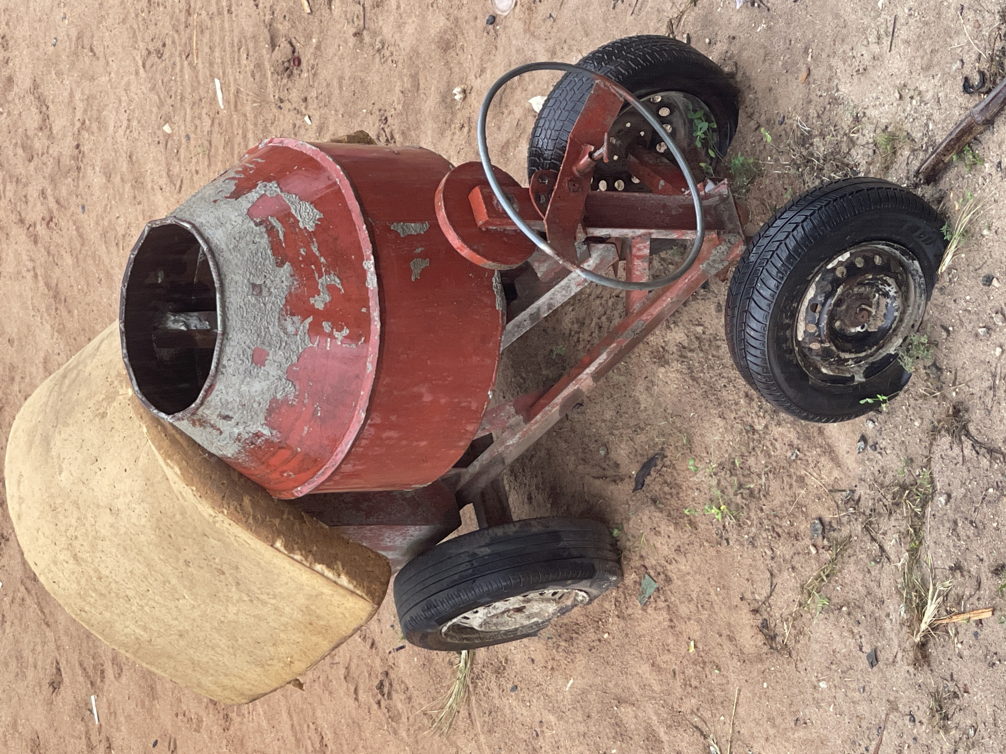A red, worn concrete mixer rests on sandy ground in Abraka, Nigeria. The mixer exhibits peeling paint and rust. It features a drum, a steering wheel-like mechanism for operation, and three tires. A tan-colored object is positioned near the drum's opening. Small patches of vegetation are scattered near one of the tires.
