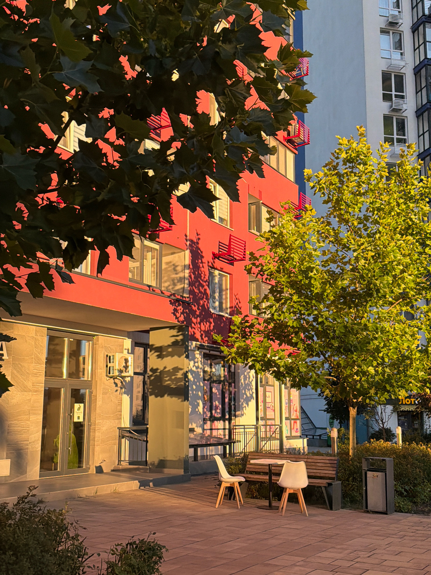 In a paved outdoor area in front of modern residential buildings in Kyiv, Ukraine, a sunlit scene unfolds. The foreground features red brick paving stones, with a small, dark green bush in the lower left corner. A paved pathway leads towards the buildings.
To the left, a building facade is painted a vibrant red. It has several windows and balconies with red railings. An air conditioning unit is visible on the lower section of the building. The entrance to this building is a glass door with a light stone-tiled frame.
To the right, a large green tree with dense foliage is bathed in golden sunlight, casting dappled shadows. Behind this tree, another modern building with a light blue and white facade rises, featuring numerous windows and white air conditioning units.
In the middle ground, a wooden bench sits beside a small table. Two white chairs with wooden legs are placed around the table, suggesting an outdoor seating area. A dark gray trash can is positioned next to the bench. The ground in this area is also paved.
The scene is illuminated by the warm, late afternoon or early morning sun, creating strong contrasts between light and shadow. The overall impression is one of urban residential architecture and a pleasant outdoor space. No people are present in the image.