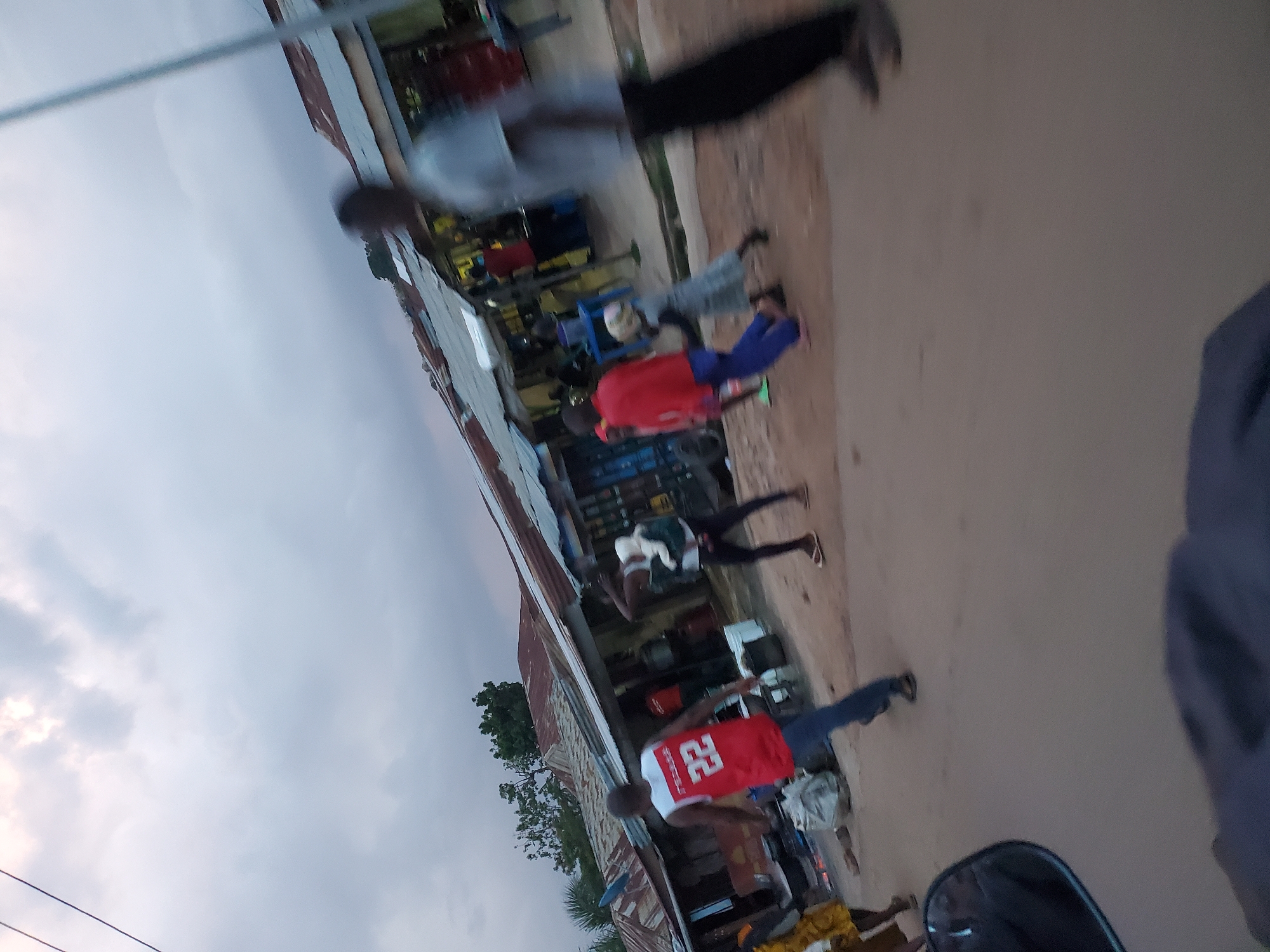 The image depicts a street scene in Jos, Nigeria, with several people walking and interacting in front of small shops with corrugated metal roofs. The sky is overcast, suggesting it might be late afternoon or evening. The ground appears to be a dirt road.
In the foreground, on the left side, a person wearing a yellow dress is partially visible. A man in a red basketball jersey with the number "22" is walking away from the camera, with a group of people behind him. One person in this group is wearing a white top and dark pants, and appears to be carrying a baby. Further to the right, a person in a red shirt and blue pants is walking. Another child in a grey dress is also present. A blurred figure of a person wearing dark pants and a grey shirt is on the far right.
The shops in the background are lined up along the street, with some appearing to be selling goods. Bags of what look like grains or other commodities are visible on the ground near the shops. The overall impression is of a bustling, everyday scene in a Nigerian city.