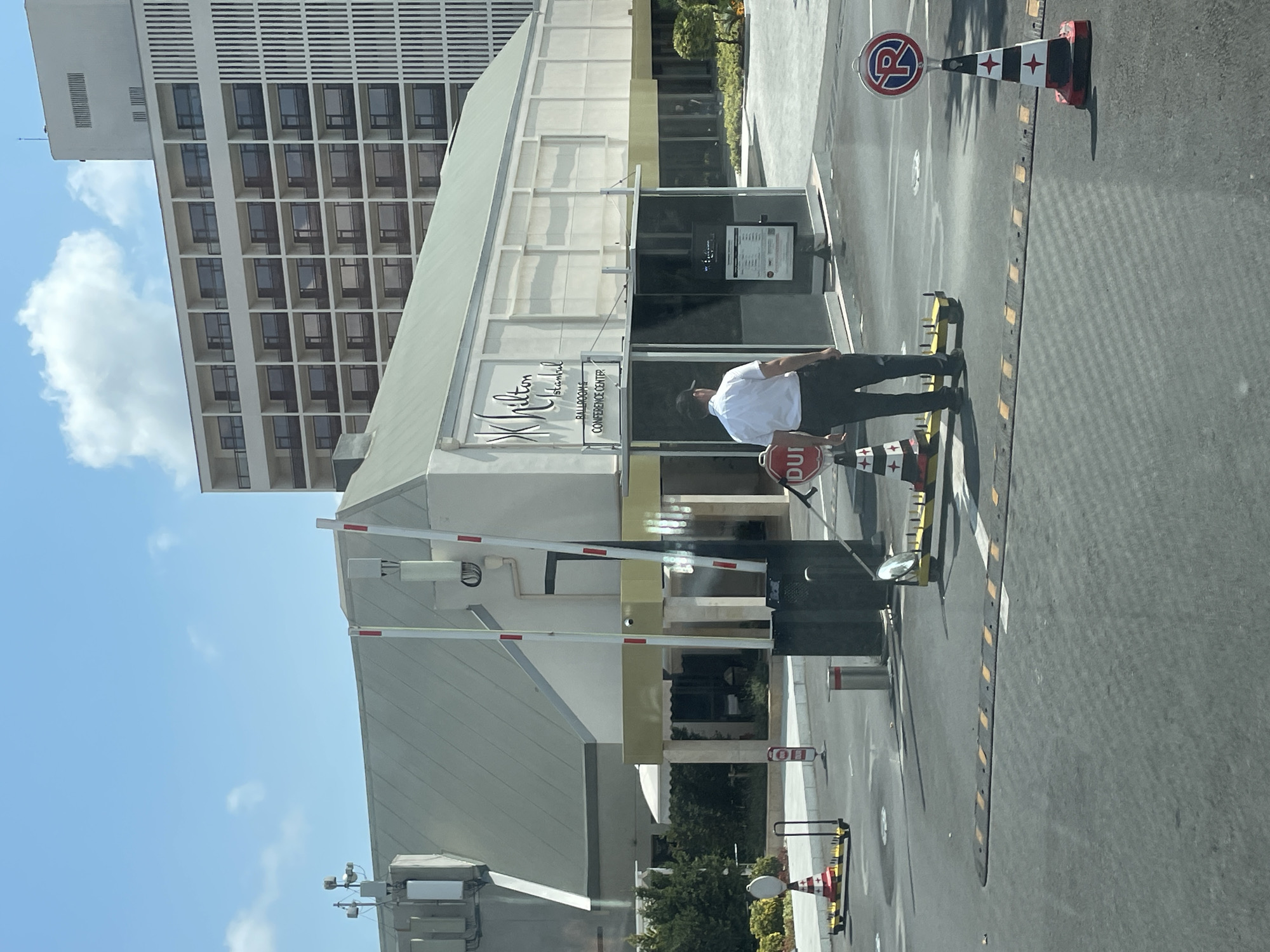 The image shows the entrance to the Hilton Istanbul Bomonti Hotel in Şişli, Turkey. In the foreground, there is a security checkpoint with a barrier arm, a security guard, and traffic calming measures on the asphalt. The security guard, a male wearing a white short-sleeved shirt and dark trousers with a baseball cap, stands near a booth. Various signs and cones are visible, including a "STOP DUI" sign, a parking sign with the letter "P", and several traffic cones. In the background, the Hilton Istanbul Bomonti Hotel complex is visible, featuring a modern building with multiple floors of balconies. The sky is clear with a few clouds. The overall scene depicts a well-maintained hotel entrance with security in place.