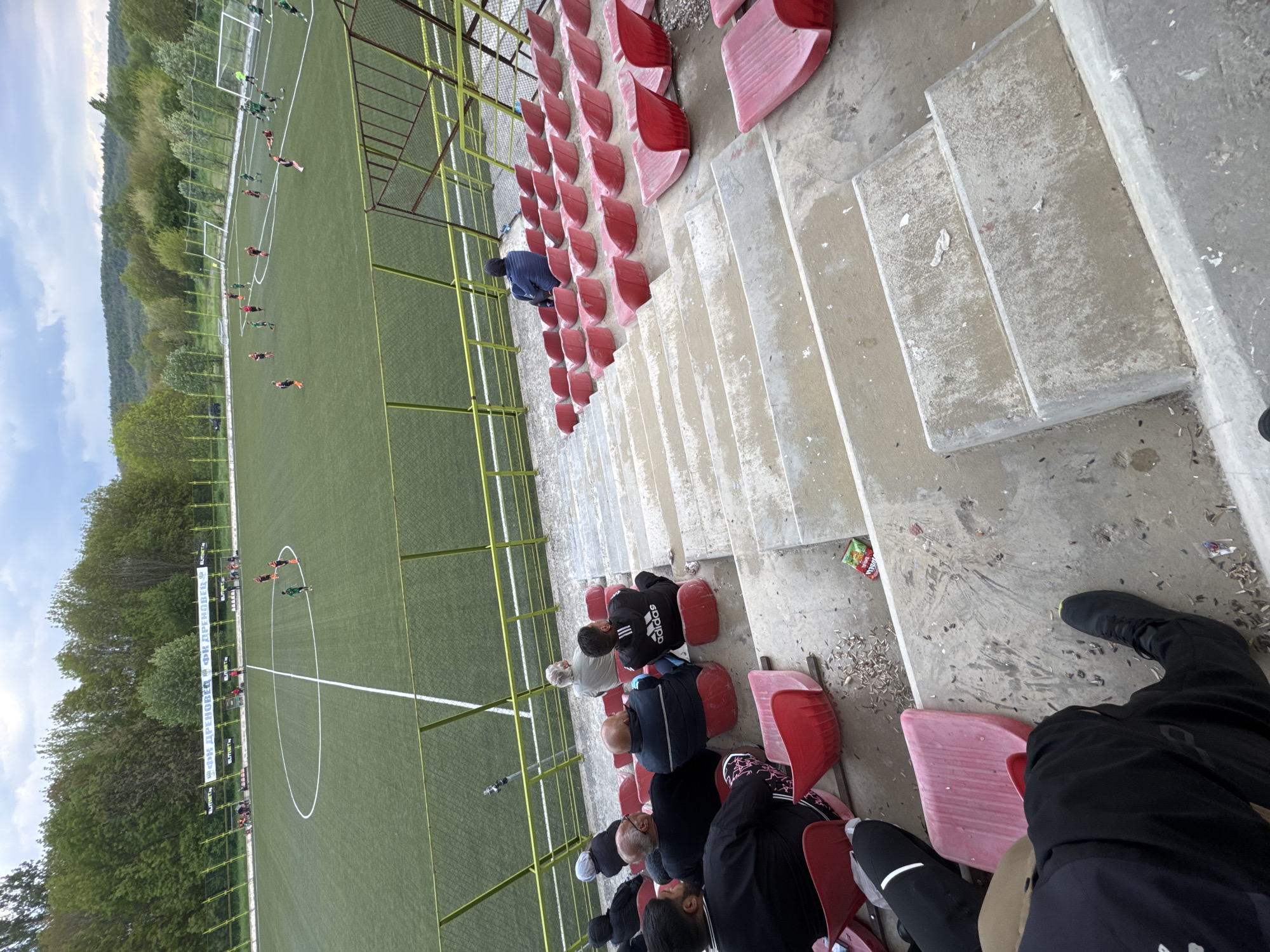View of a football field in Drenovets, Bulgaria, from a seating area. The field is green artificial turf, with white lines marking the playing area. Players in green and red uniforms are visible on the field. A fence separates the field from the seating area. The seating area consists of concrete steps and red plastic seats. Several people are seated, watching the game. The ground is littered with debris. The background features trees and a cloudy sky.