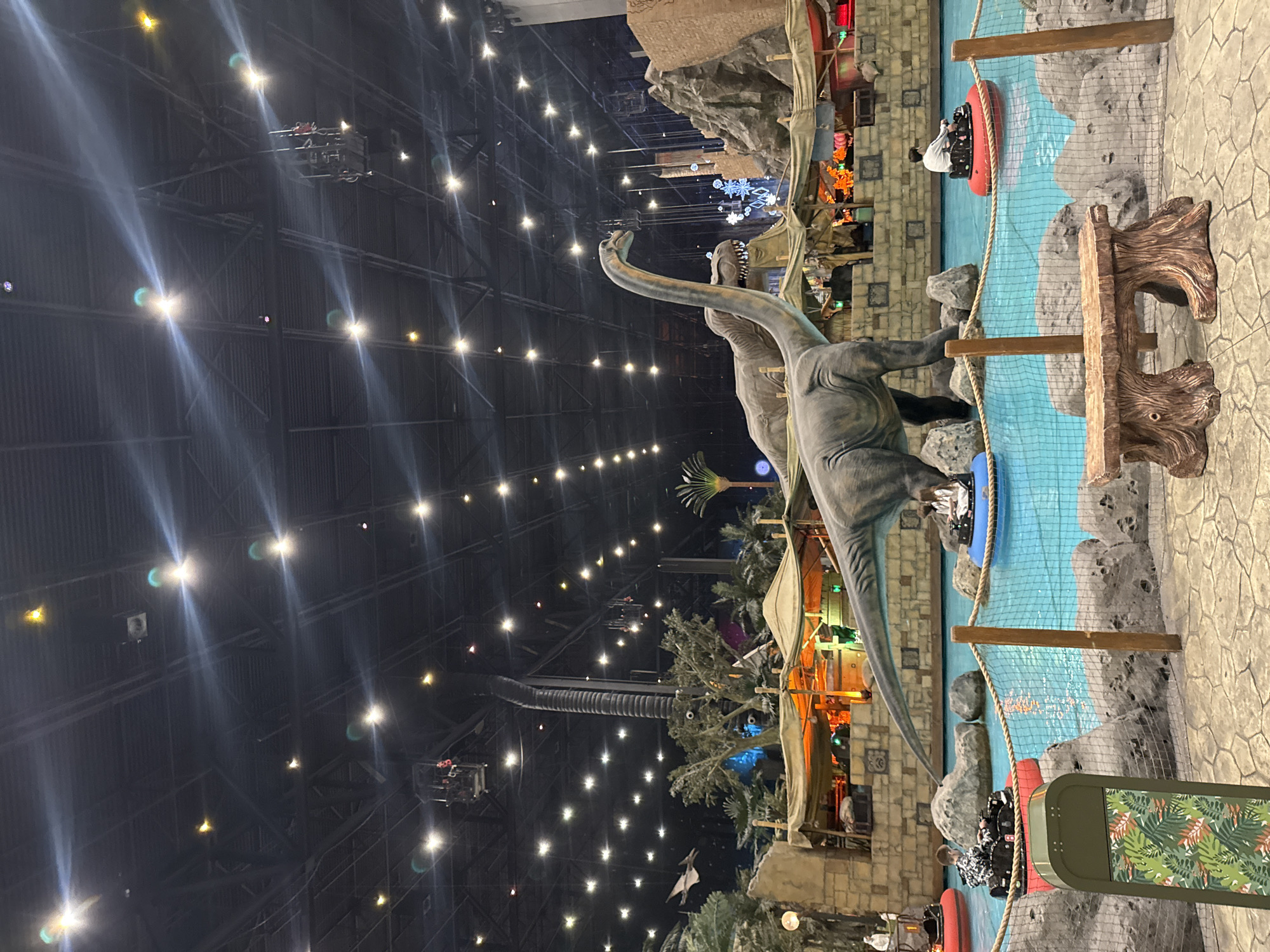 This image depicts a vibrant, dinosaur-themed indoor amusement park or entertainment center, likely situated in Moscow, Russia. The central focus is a large, detailed statue of a long-necked dinosaur, resembling a sauropod, alongside another theropod-like dinosaur statue partially visible behind it. These statues are positioned next to a winding artificial river or canal, where visitors are enjoying a water ride in red inflatable boats.
Several individuals can be seen in these boats, navigating the clear blue water. One person wearing a white top and another in dark attire are clearly visible in separate boats. The water feature is bordered by faux rock formations, a safety net suspended between wooden posts, and textured pathways. The setting is elaborately decorated to mimic a prehistoric jungle environment, complete with artificial palm trees and dense green foliage. In the background, structures resembling ancient ruins or jungle huts are illuminated with warm lights, contributing to the immersive atmosphere.
The overarching ceiling of the facility is exceptionally high and dark, studded with numerous bright, warm-toned lights that create prominent light streaks and flares, suggesting an evening or nighttime operational setting under artificial illumination. Metal girders, support structures, and industrial ventilation ducts are visible high above, revealing the vast scale of the enclosed space. A decorative panel with a tropical leaf pattern in green and white is visible in the foreground, aligning with the themed environment. The scene portrays a lively and engaging recreational activity within a highly themed indoor setting. No discernible text is visible in the image.