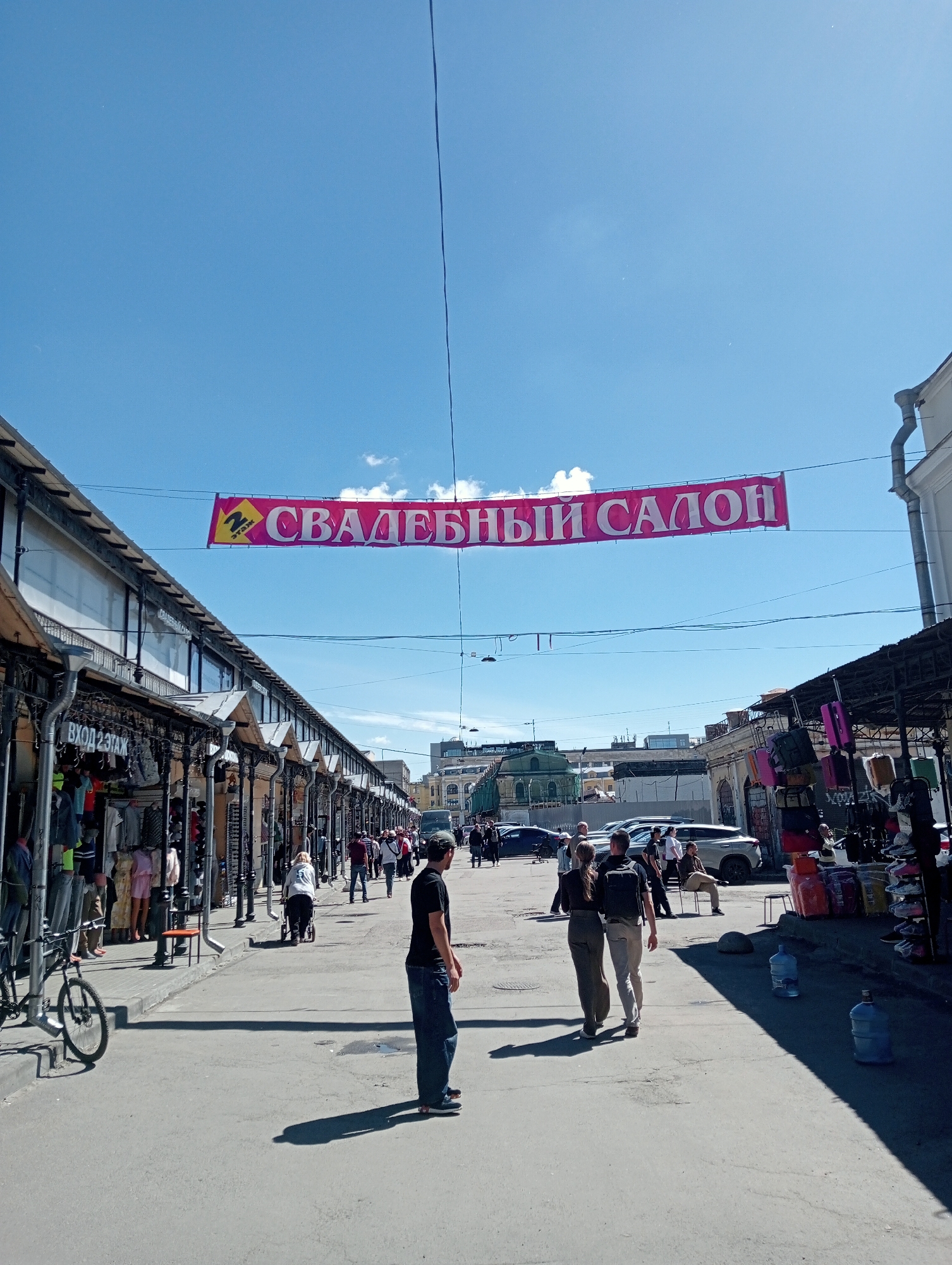 The image depicts a street market scene in Saint Petersburg, Russia, under a bright blue sky with scattered clouds. A large pink banner with yellow text, reading "СВАДЕБНЫЙ САЛОН" (Wedding Salon), is strung across the street overhead. The market features rows of stalls with awnings, some displaying clothing and other goods. People are walking along the street, browsing the stalls, and interacting. In the foreground, a man in a black t-shirt and jeans stands with his back to the camera, casting a long shadow. To the left, a bicycle is parked near a stall. The street is paved, and cars are visible in the background. The overall atmosphere suggests a bustling daytime market.