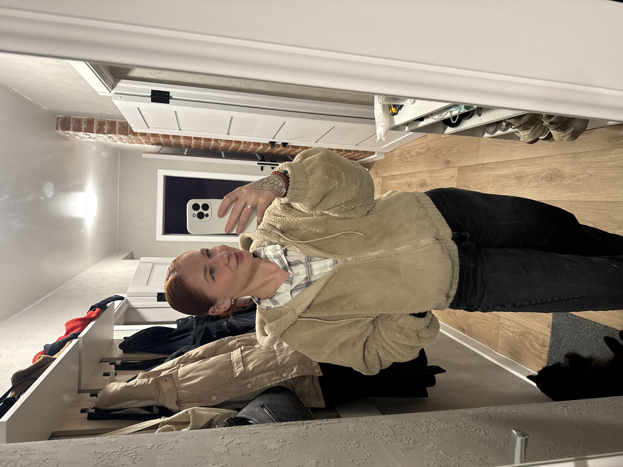 A red-haired woman is taking a selfie in what appears to be a hallway. She is wearing a beige fleece jacket over a plaid shirt and black jeans. The hallway contains a coat rack with various jackets and bags on it. The floor is made of wood, and there's a glimpse of a black cat on the floor. There is also a shelf above the hallway with shoes on it. A brick wall is visible through a doorway in the background. The photo does not provide information about the city of Rostov-on-Don, Russia.