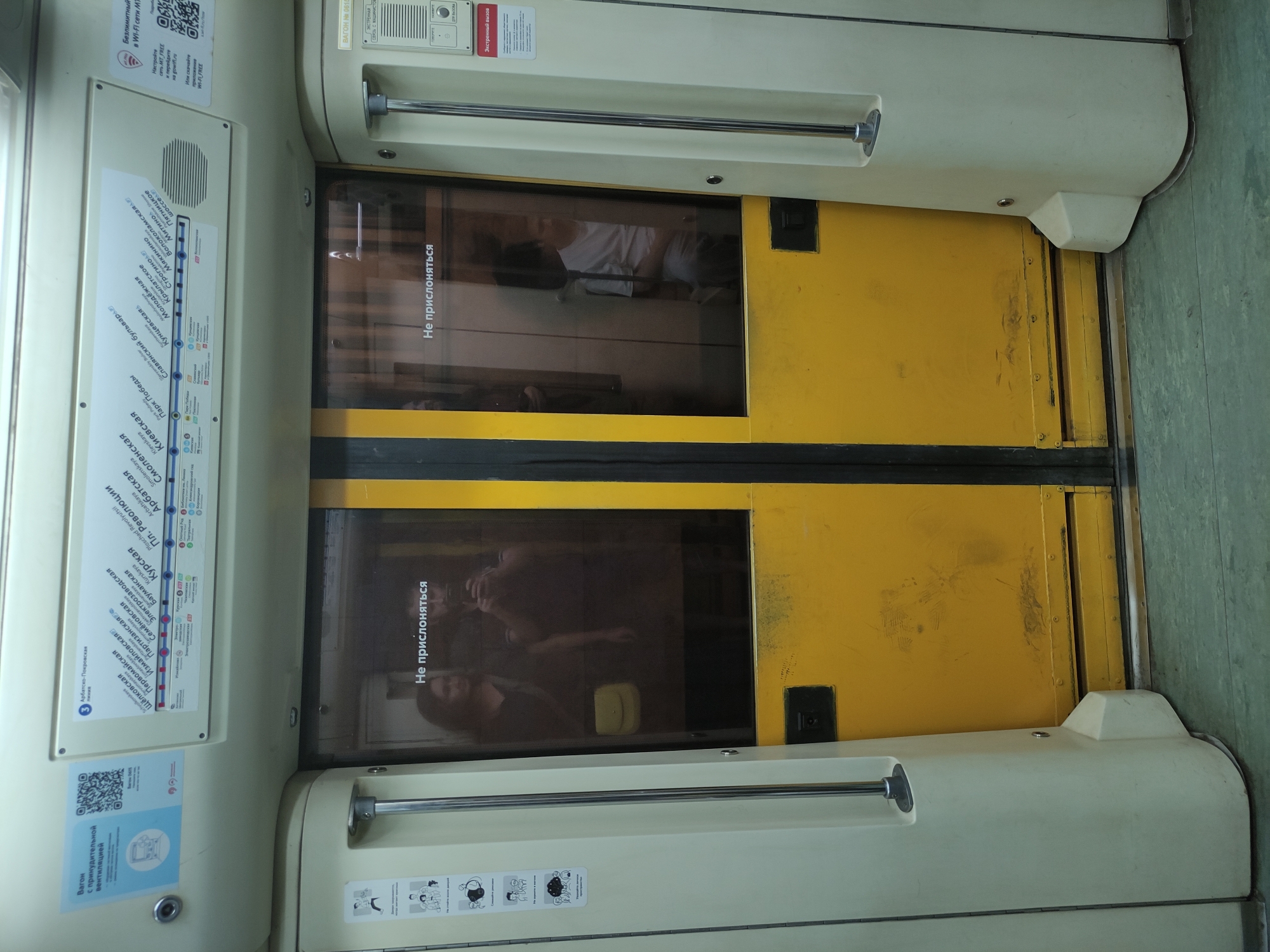 The image shows the interior of a Moscow Metro train, specifically the doors leading to another carriage. The doors are yellow and have rectangular windows with a warning sign in Russian that translates to "Do not lean." The doors have horizontal grab bars. The inside of the train is white and beige with stickers showing the metro line route. The metro map sticker shows the Arbatsko-Pokrovskaya line, which is line number 3. Stops like "Shchelkovskaya," "Kievskaya" and "Slavyansky Boulevard" are visible on the map, placing the location within the Moscow Metro system. Other stickers indicate the presence of Wi-Fi and provide emergency contact information. Reflections in the window suggest there are people in the other carriage.