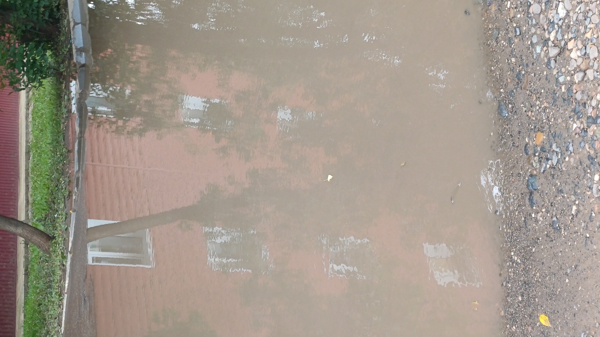 The image depicts a muddy puddle in front of a building, possibly in Izhevsk, Russia. The puddle reflects the facade of the building, which appears to be made of reddish-brown brick. Several windows are visible in the reflection, as well as the reflection of a tree. The ground around the puddle is covered in gravel and dirt. A small fish is visible swimming in the puddle. The scene suggests an outdoor environment, likely a street or a courtyard, after a period of rain.