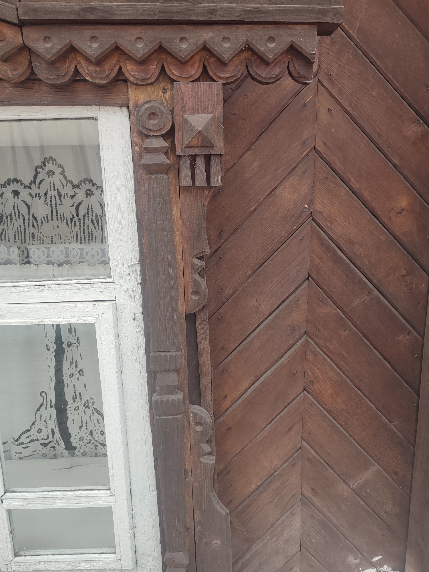An eye-level close-up shot reveals a section of a wooden structure, possibly an old building, located in Sarapul, Russia. The structure is primarily constructed from brown wooden planks arranged in a chevron pattern. Adjacent to this wooden wall is a white-framed window with a lace curtain visible through the glass. The window frame features intricate wooden carvings along its vertical edge, showcasing detailed patterns and shapes. The wooden wall also has similarly detailed carvings where it meets the window. The scene is well-lit, highlighting the texture and detail of the wood and carvings.