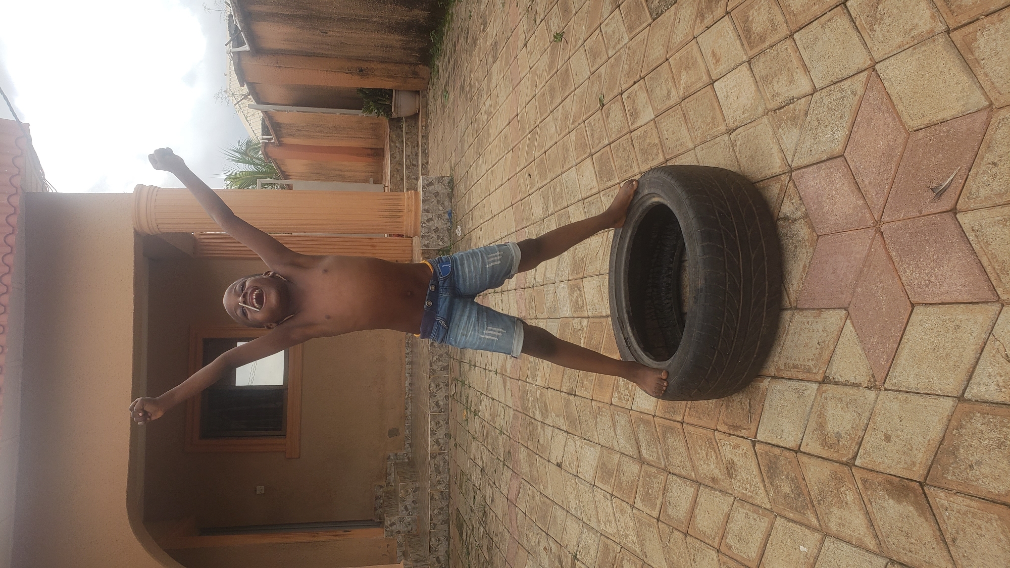 A young Black male, shirtless and wearing denim shorts, stands with his arms raised in celebration while balanced on a tire in an outdoor courtyard. The courtyard is paved with interlocking square and star-shaped tiles in shades of tan and reddish-brown. Behind the boy, a light-brown building with a covered porch supported by a column is visible. The building has an open window with a dark interior. The scene is set in Sabon Tasha, Nigeria.