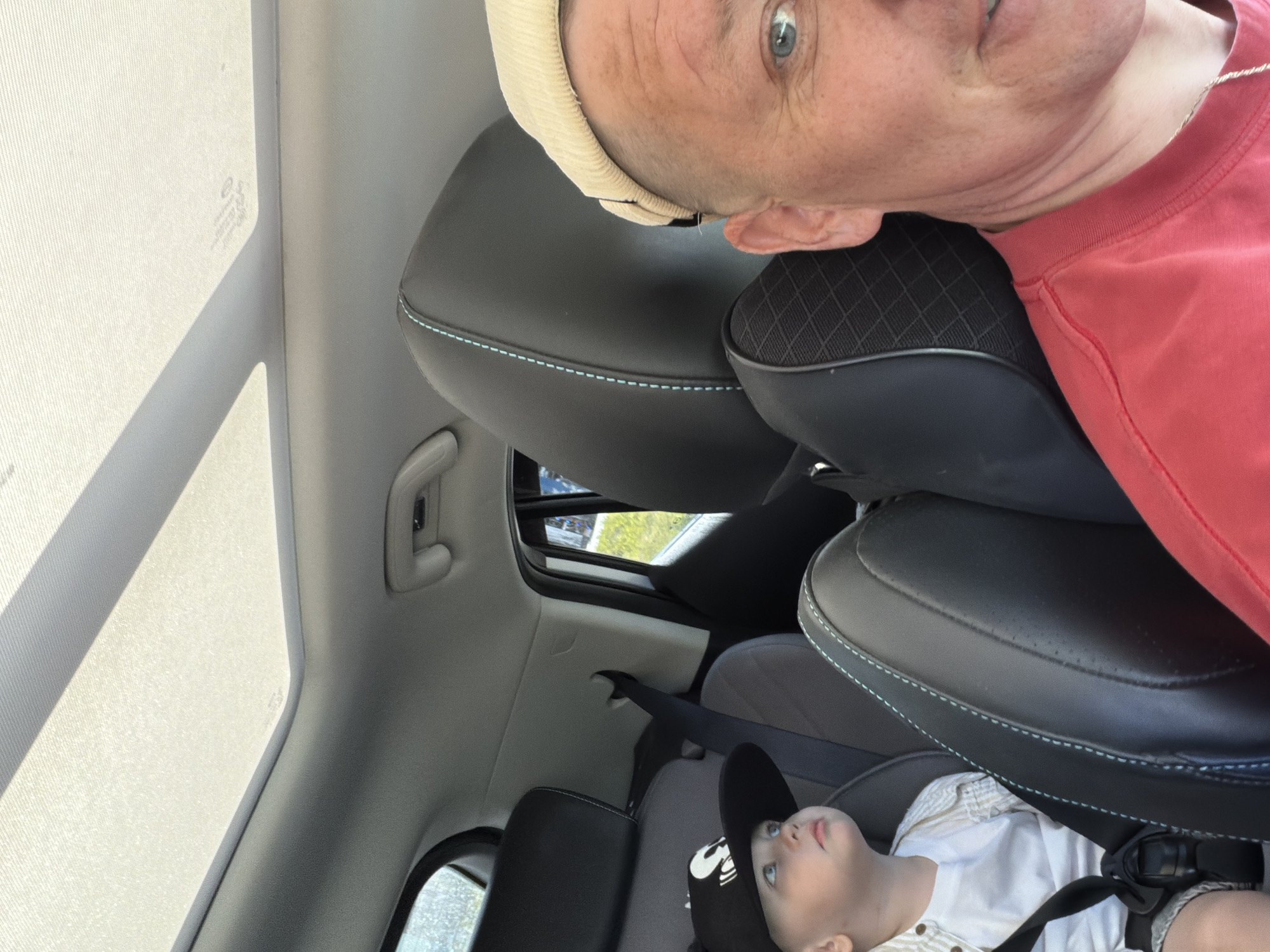An outdoor, daytime, eye-level selfie captures a man in the foreground, partially visible from the chest up, wearing a red t-shirt and a light-colored beanie. His blue eyes are looking towards the camera. Behind him, a young child is visible in a car seat, wearing a black baseball cap with a white logo and a white shirt. The child is looking upwards and to the left. The background shows the interior of a car, including black leather seats with light blue stitching and a sunroof. The image appears to have been taken while the car is in motion, as suggested by the slight blur in the background and the way light streams through the sunroof. The image does not contain any identifiable landmarks or features that would indicate the specific location within the city of Chaikovsky, Russia.