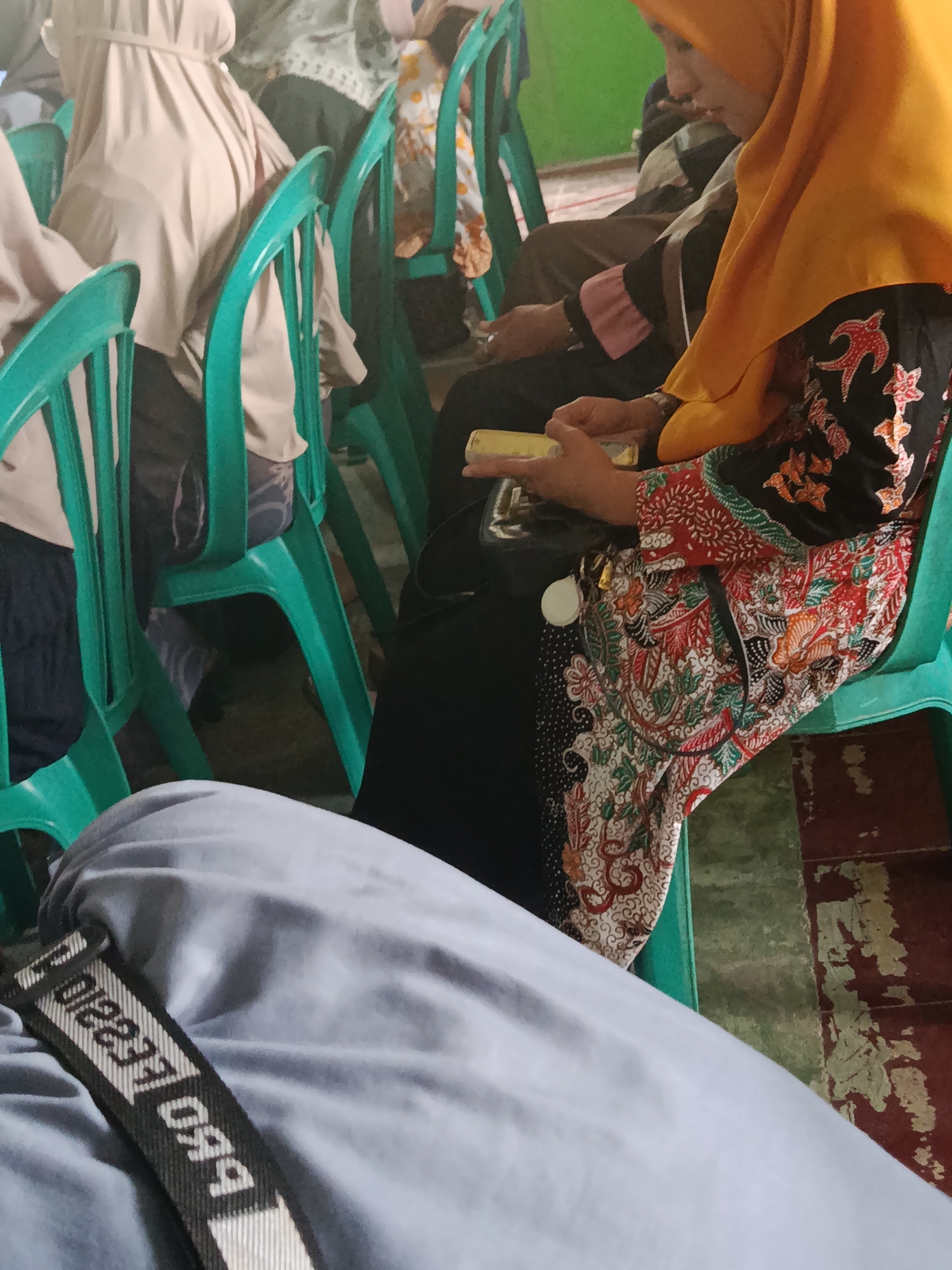 The image depicts an indoor scene with several individuals seated on bright green plastic chairs. In the foreground, a person wearing light blue pants and a black strap with white text is visible. The strap has the word "LEGA" printed on it. Behind this person, a woman wearing an orange hijab and a patterned black and red batik dress is seated. She is holding a yellow object in her hands. To the left, another person is partially visible, wearing a light beige hijab. The chairs are arranged in rows, suggesting a gathering or event. The environment appears to be a room with green walls and a floor made of red and green tiles, possibly in the village of Kawunganten Lor, Indonesia.