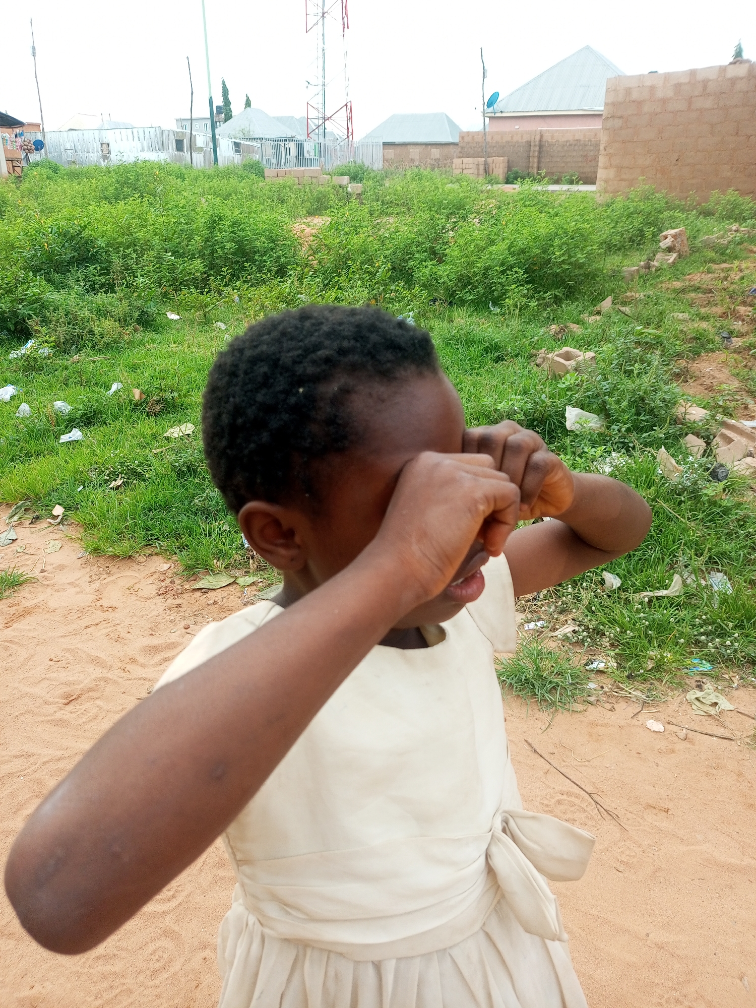 A young Black girl, likely a child, is the central subject of this photograph. She is wearing a light-colored dress with a sash tied at her waist. Her hands are clasped over her eyes, suggesting she might be crying, playing a game of peek-a-boo, or simply shielding her eyes from the sun. Her dark, short hair is visible around her face.
The setting appears to be an outdoor environment, possibly in Tempure, Nigeria, given the context. The ground is sandy and dry, with patches of green grass and weeds growing sporadically. Some scattered white plastic waste is visible on the ground, indicating a potentially unkempt area. In the background, there are several buildings under construction or recently built, made of brick and corrugated metal. A prominent cell tower with a red and white structure stands in the mid-ground. The sky is overcast, suggesting a cloudy day. The overall mood is ambiguous, with the girl's hidden face leaving room for interpretation of her emotions. No visible text is present in the image.