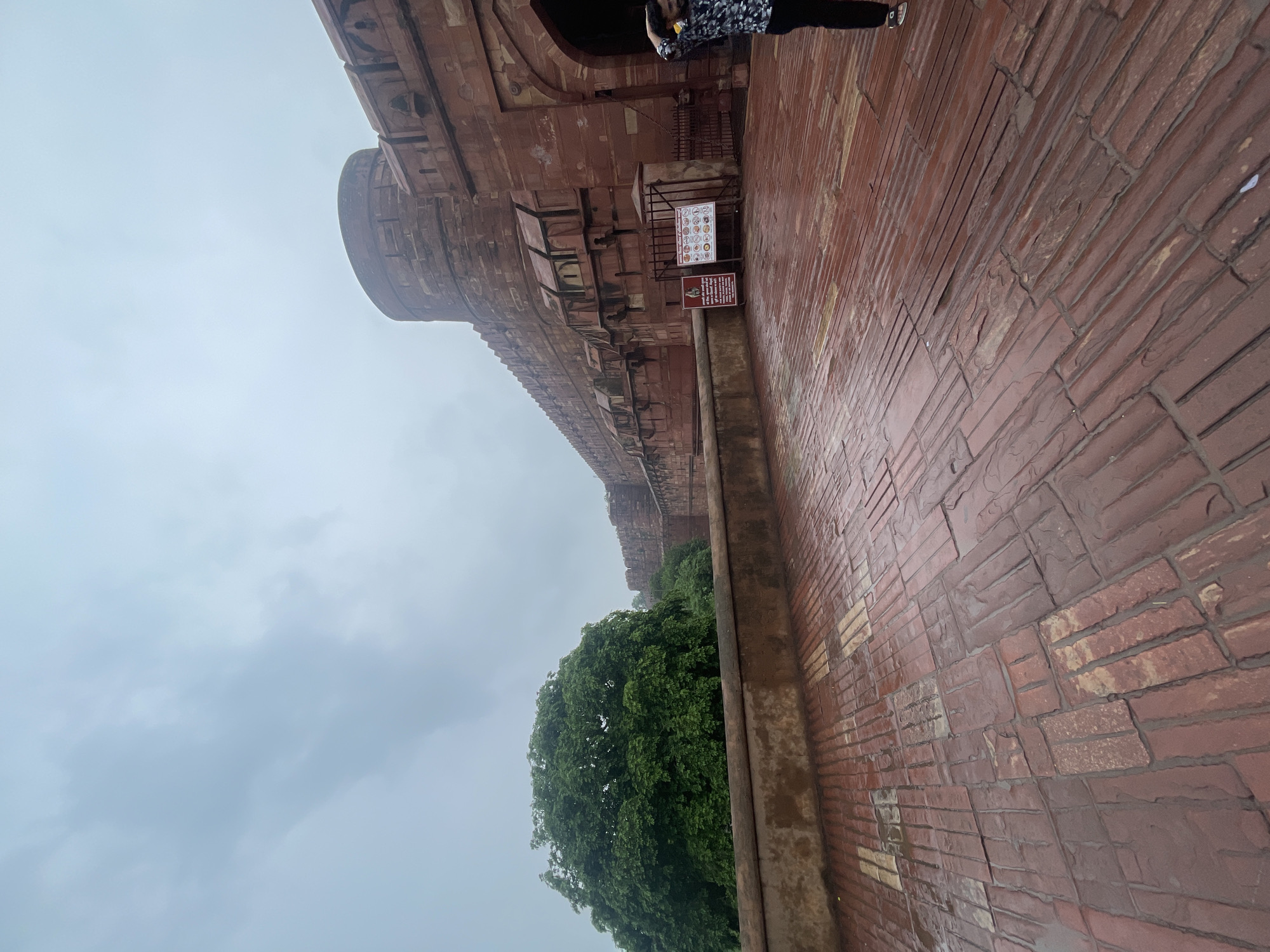 A low-angle, wide shot captures a portion of the Agra Fort in Agra, India, on an overcast day. The foreground is dominated by a wet, red brick courtyard, with the bricks laid in a staggered pattern, reflecting the dim light. To the left, a low, tan-colored wall runs along the edge of the courtyard, behind which a lush, dense green tree stands tall. Beyond the tree, the imposing red sandstone walls of the fort stretch into the distance, featuring a large, rounded bastion and smaller crenelated sections. Multiple arched openings are visible in the fort's facade. Two informational signs, one with text and an image of a person, and another with a grid of colorful icons, are positioned near the wall. In the far right of the image, a person wearing a dark, patterned shirt and black pants stands with their right hand behind their head, looking towards the fort. The sky is a pale, overcast gray.