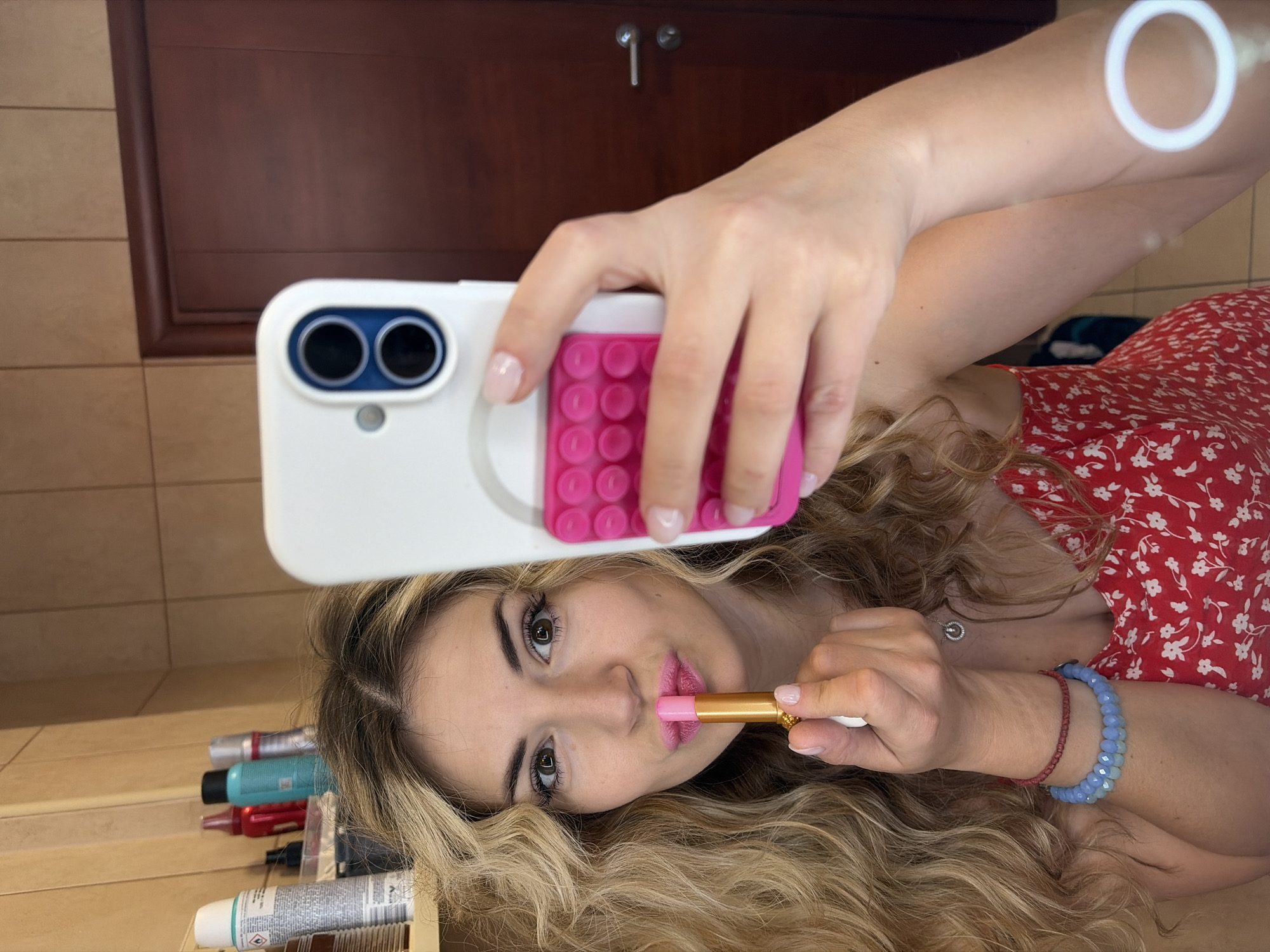 A selfie shows a woman with long, wavy blonde hair applying pink lipstick. She wears a red floral dress. She is holding a white smartphone with a pink, possibly bubble-textured grip on the back. The reflection suggests the photo is being taken in a bathroom, evidenced by the vanity, cabinet, and toiletries. The location is possibly in Budva, Montenegro (context provided).