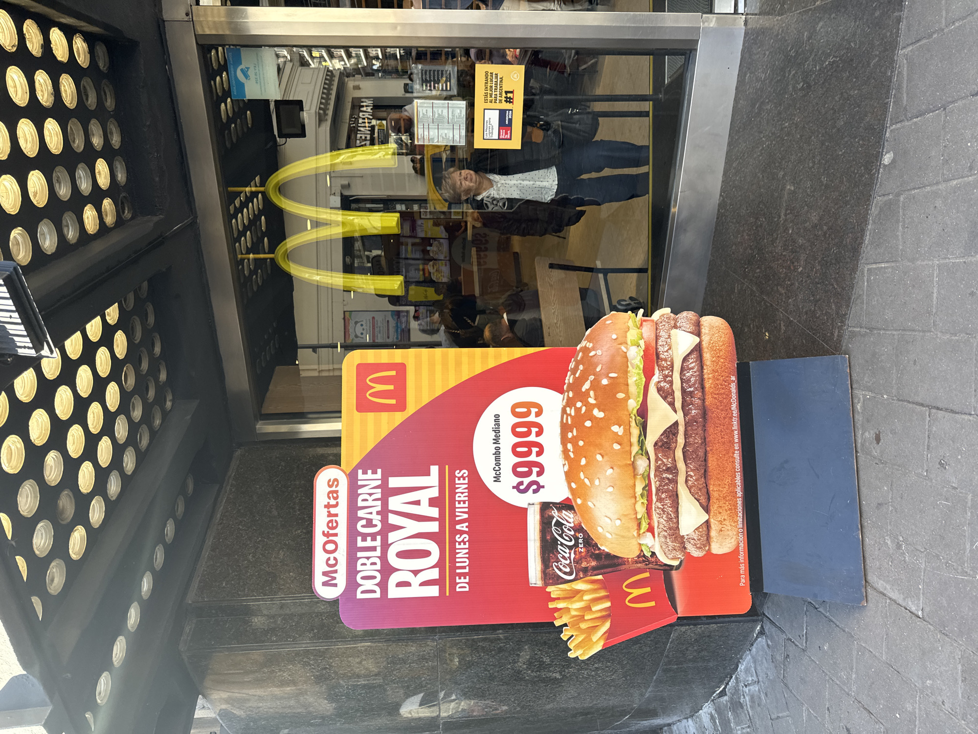The image shows the entrance of a McDonald's restaurant in Buenos Aires, Argentina. A promotional sign for a "Doble Carne Royal" meal deal costing $9999 Argentine pesos stands in front of the entrance. A woman is reflected in the glass door, standing inside the restaurant. The interior has tables and chairs, with additional signs and branding visible. The entrance is marked with the golden McDonald's arches. A sign on the door reads "ESTÁS ENTRANDO AL MEJOR LUGAR PARA TRABAJAR DE ARGENTINA." A perforated metal canopy shelters the entrance.