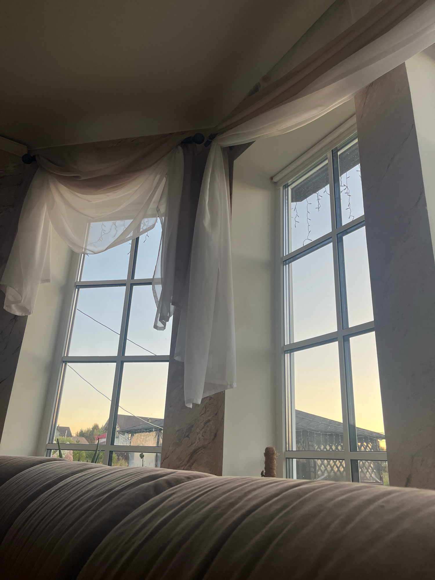 The image depicts an interior view of a room with two large windows, likely in a residential setting in Vladimirovka, Russia. A portion of a couch is visible in the foreground, covered in a textured beige fabric. The windows are adorned with sheer, draped curtains in neutral tones, creating a soft, diffused light. Through the windows, an outdoor scene is visible, characterized by the warm hues of a sunset sky. The exterior includes rooftops, trees, and a fence, suggesting a suburban or rural environment. One window has what appears to be decorative string lights hanging from the top. The overall impression is one of a quiet, domestic space bathed in the gentle light of dusk.